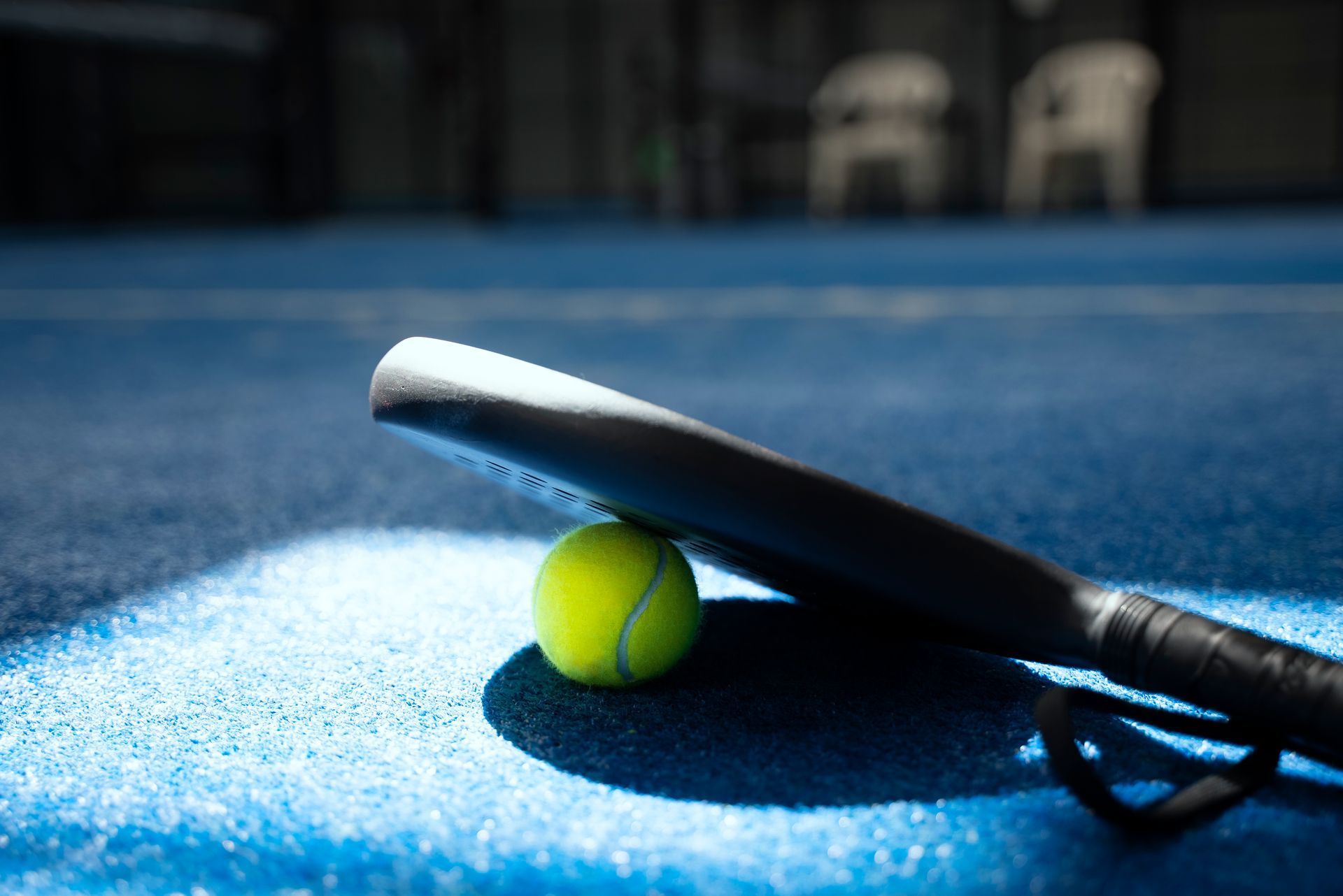 Tennis racket and ball on a blue court, with sunlight casting a shadow