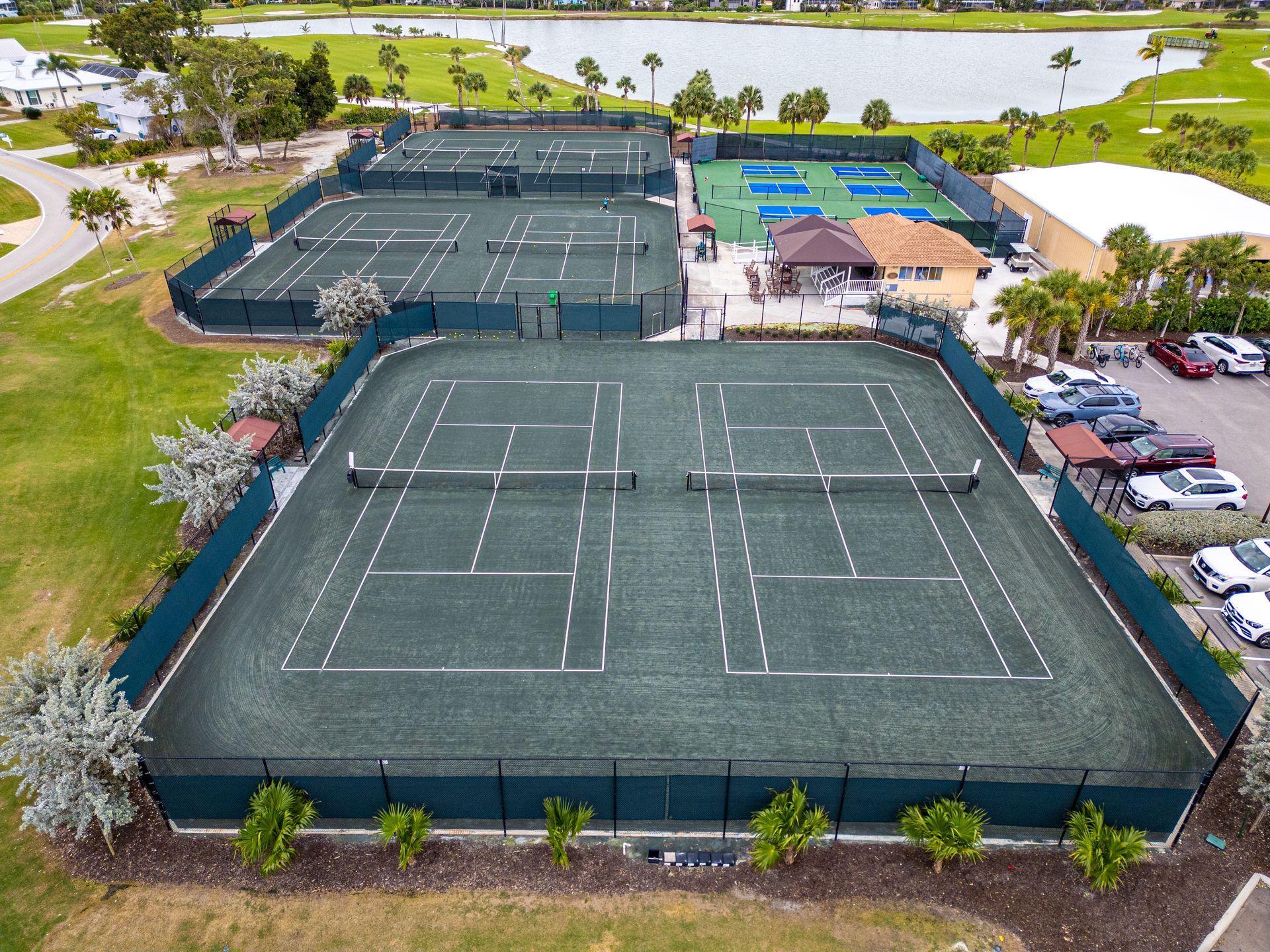 Aerial view of multiple tennis courts surrounded by fencing and parked cars near a lake.