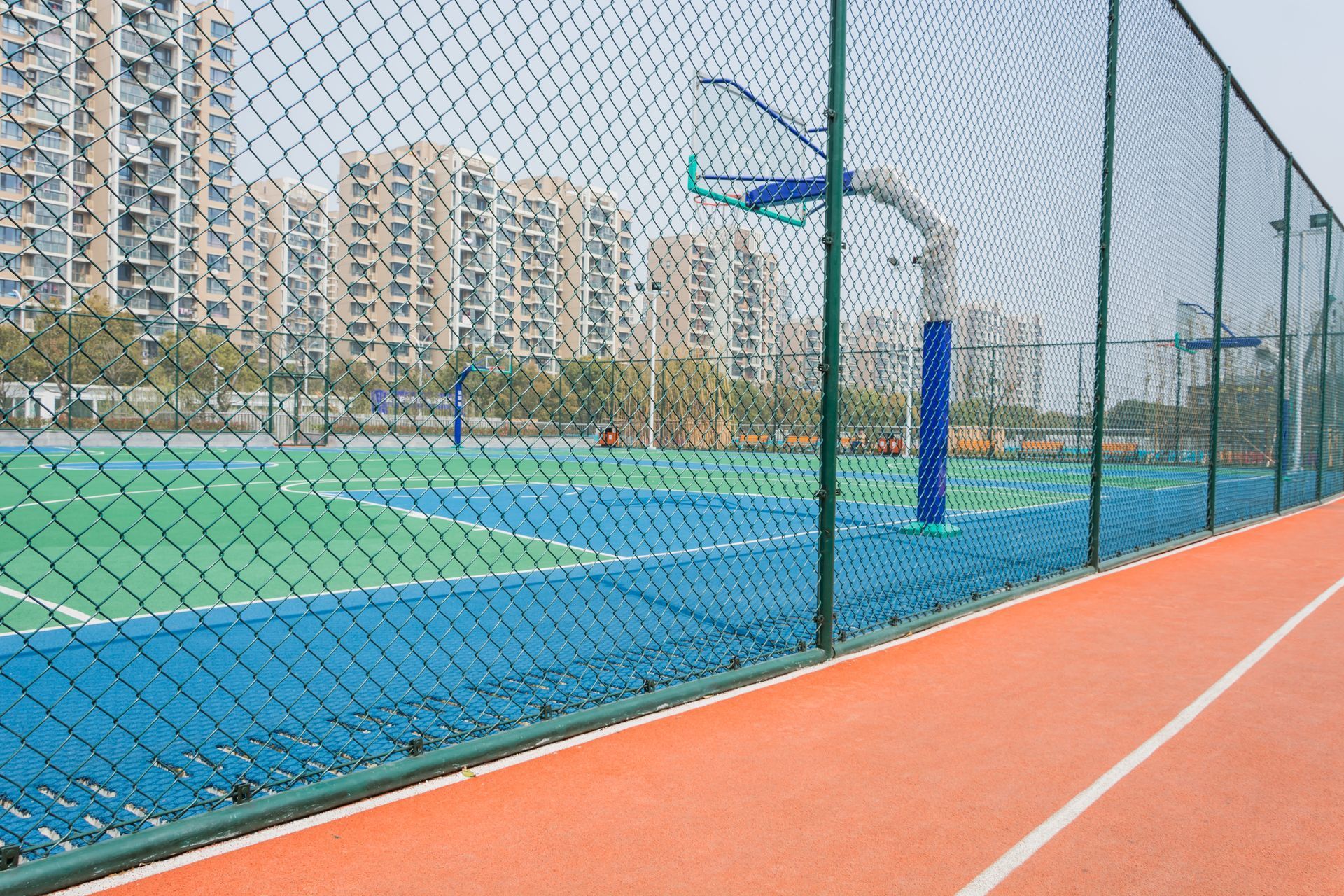 Outdoor sports facilities with a blue and green surface, netted fence, and apartment buildings in the background