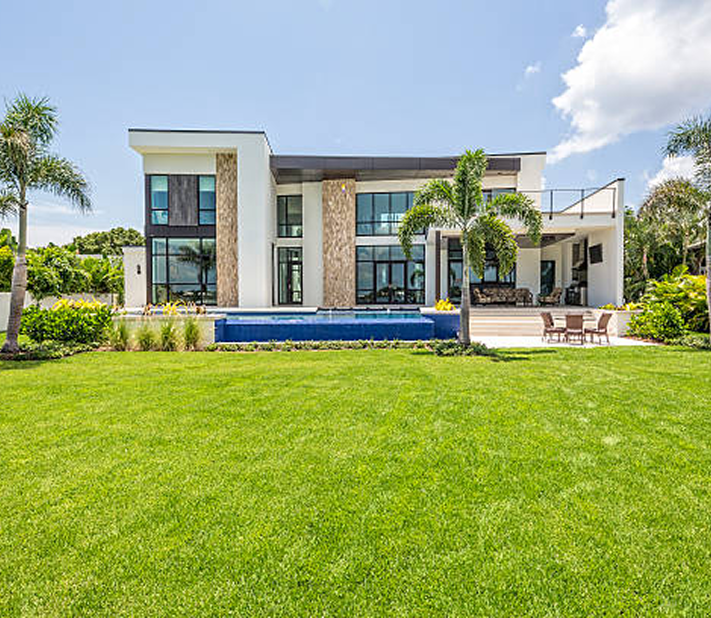 Modern white house with a large lawn, blue pool, and palm trees under a bright sky