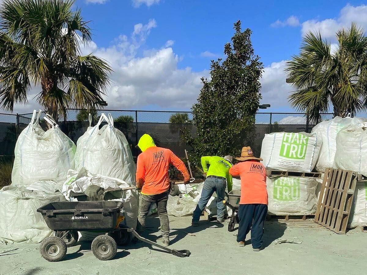 Workers loading large white bags outdoors beside a cart under palm trees and blue sky