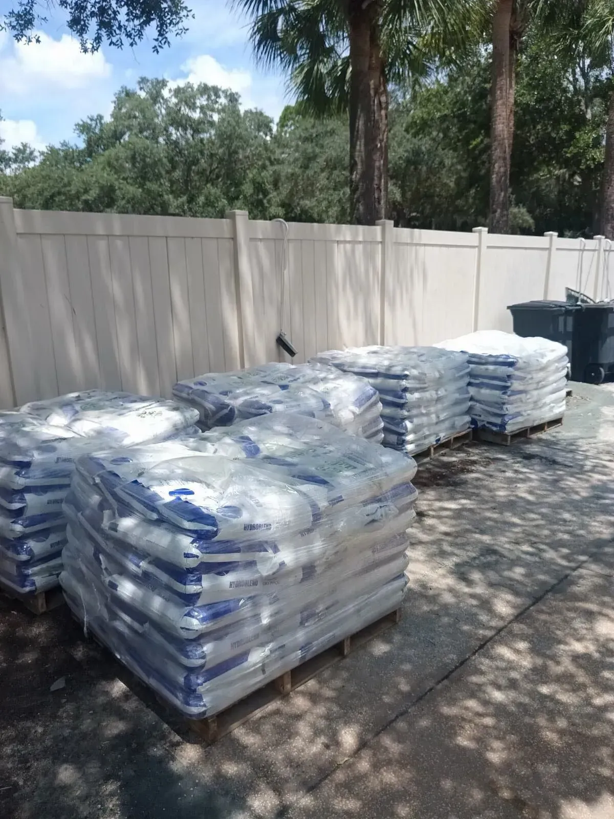 Stacks of bagged materials on pallets in a shaded outdoor yard beside a fence.