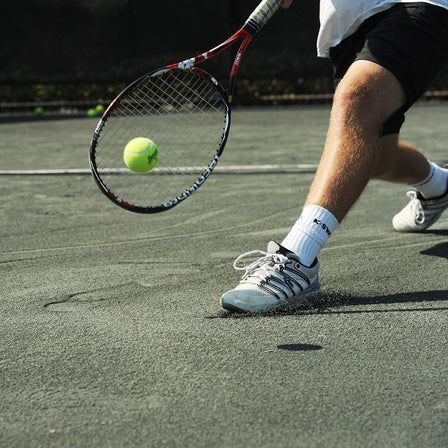 Tennis player lunging for a yellow ball on an outdoor court, racket extended low.