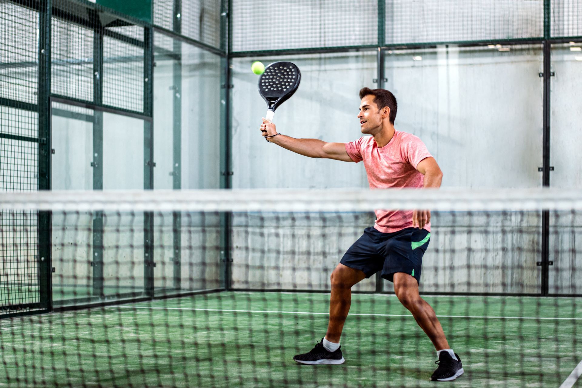 Man playing paddle tennis indoors, reaching for a ball near the net.
