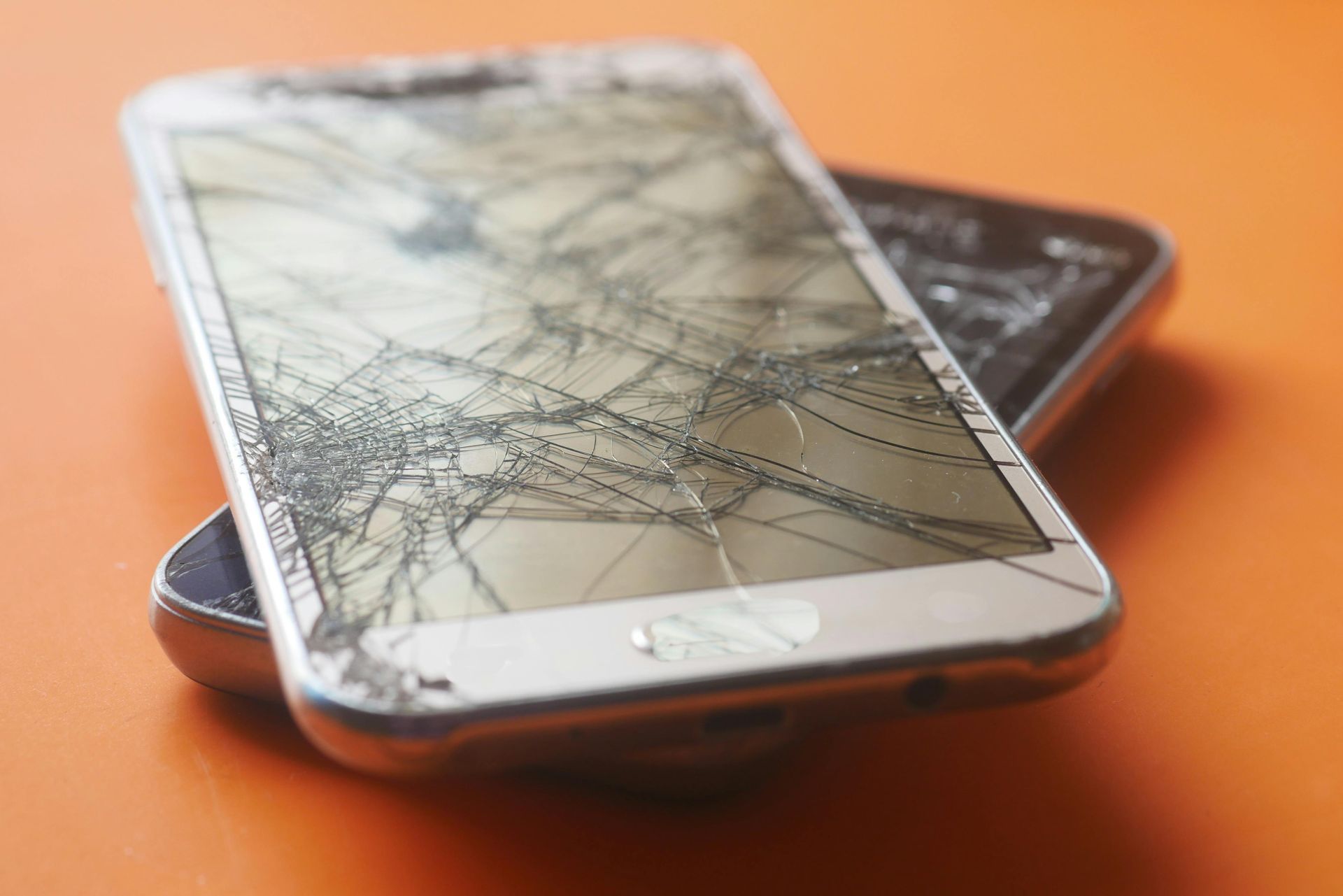 Cracked smartphone screen on an orange surface, with another phone partially underneath.
