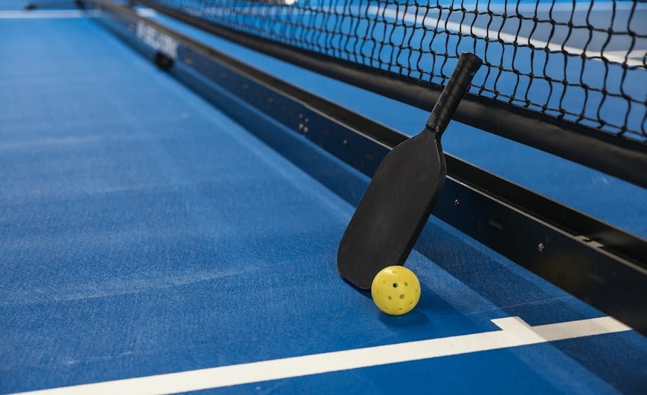 Paddle and yellow ball on a blue pickleball court near the net