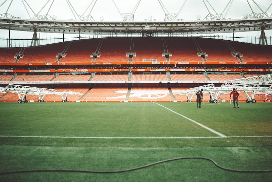 Empty soccer stadium with green pitch and red seating, grounds crew working near midfield