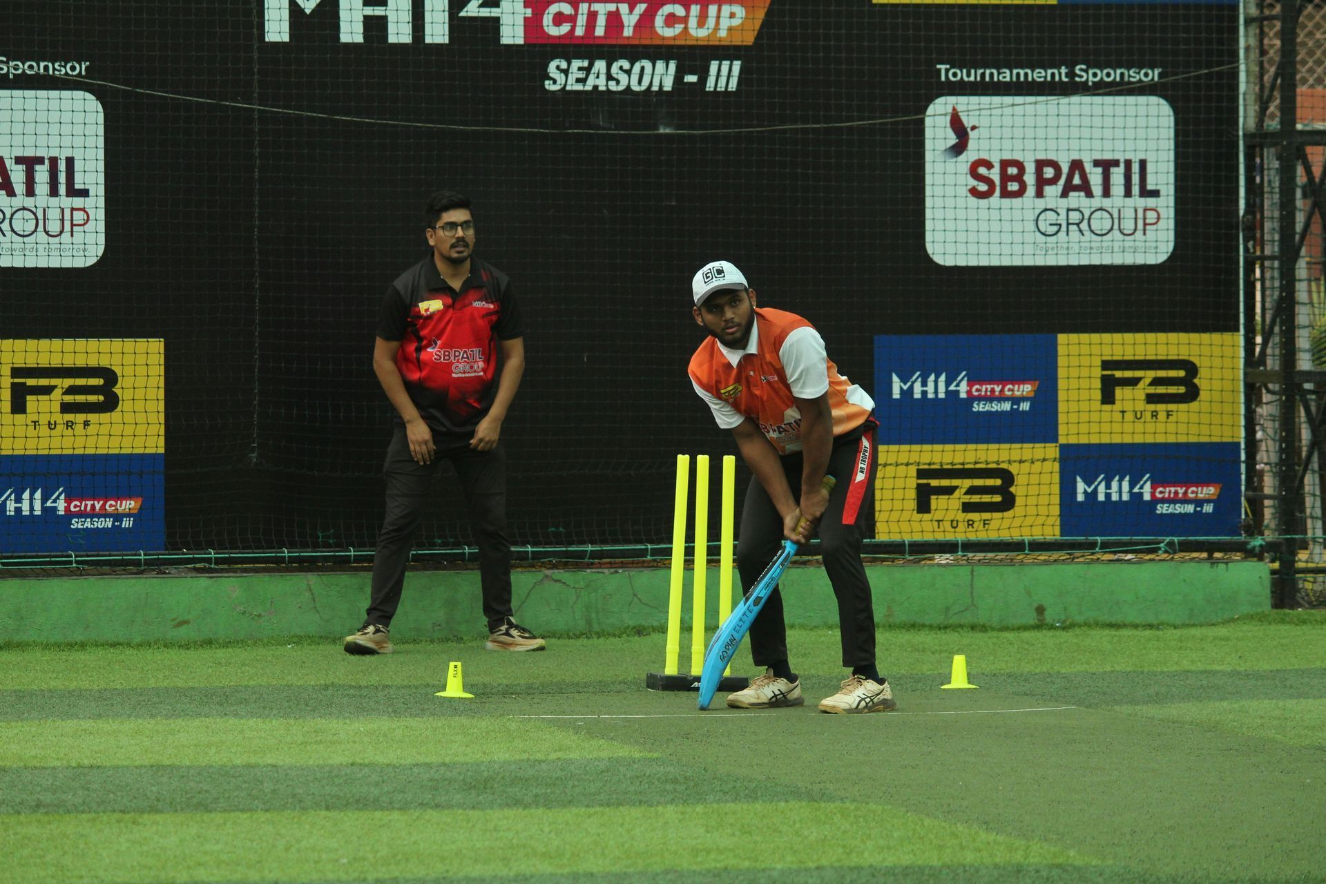 Two players on a cricket pitch, one batting with a blue bat under stadium lights.