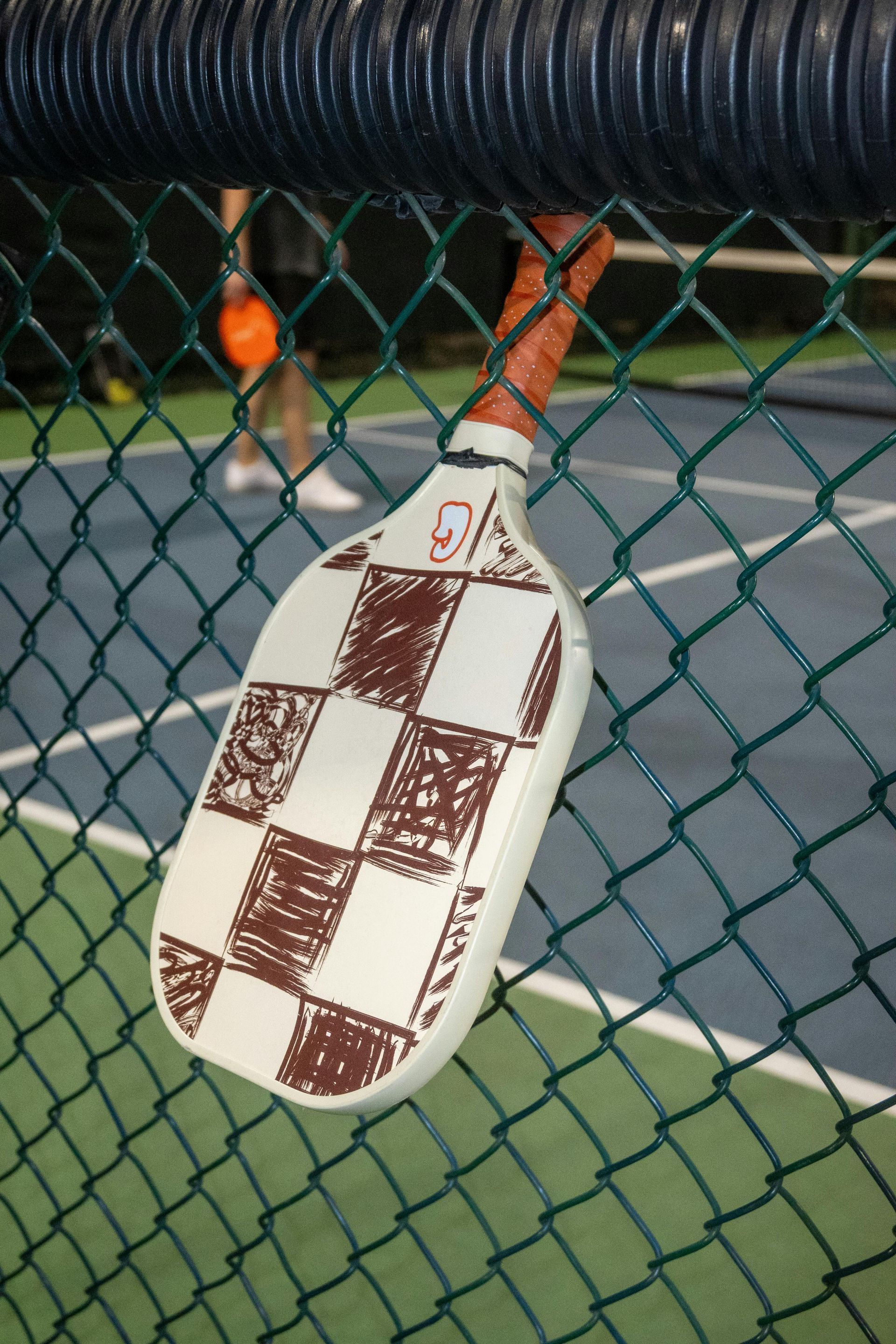Papier-mâché bat with brown checkerboard pattern hanging on a chain-link fence at a tennis court