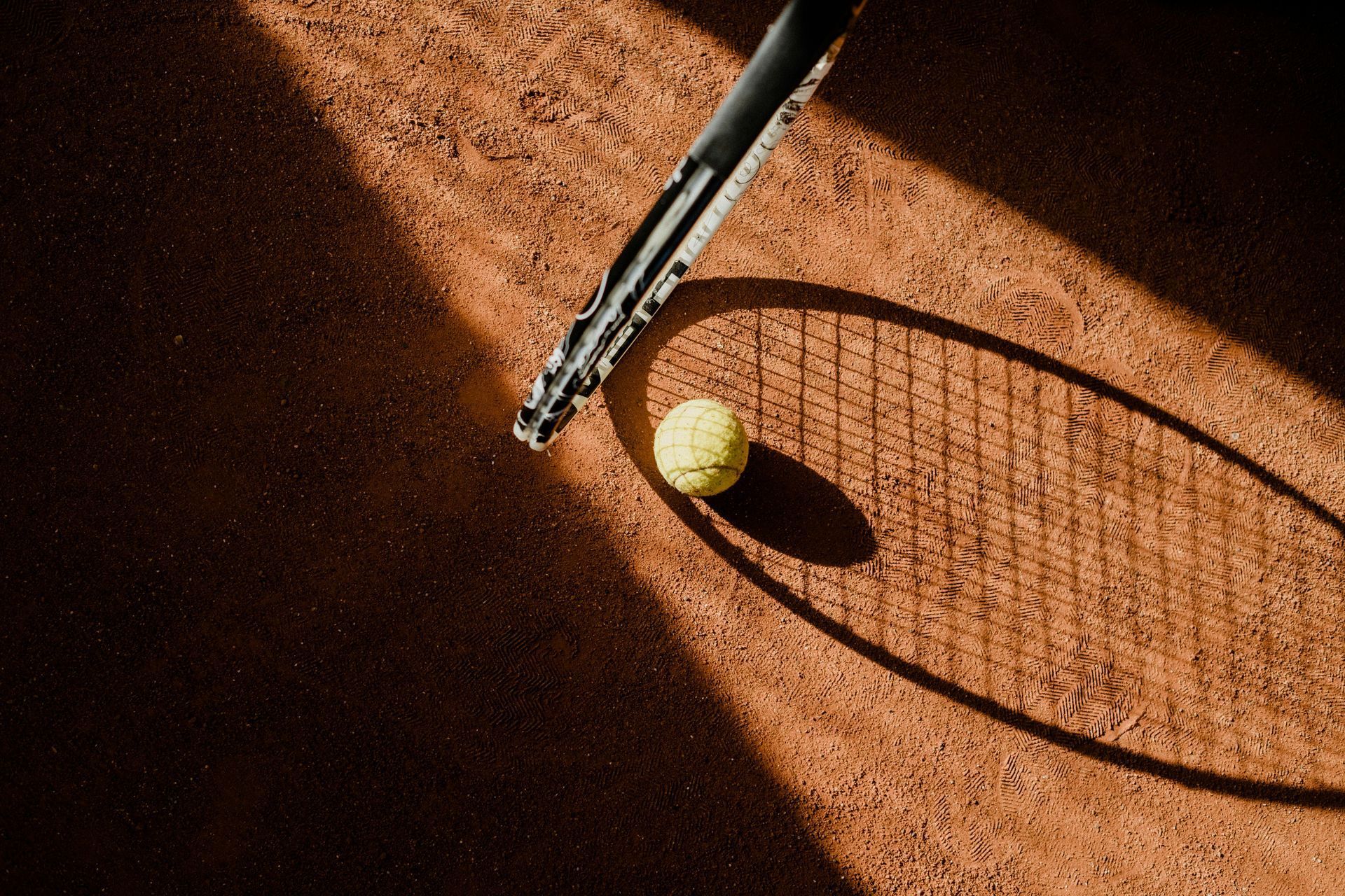 Tennis ball and racket on a clay court with sharp sunlight and shadow