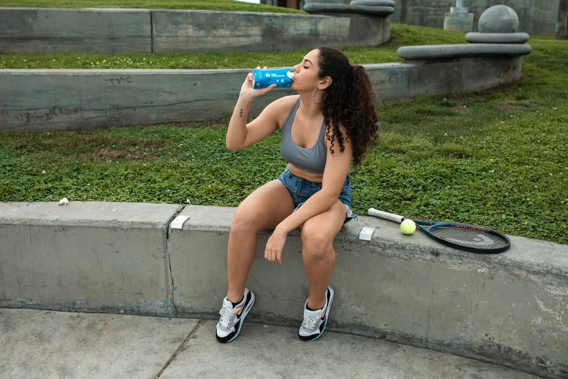 Woman in blue sportswear sits on a curb drinking water beside a tennis racket and ball.