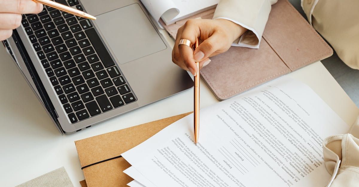 Hand holding a pencil over documents beside a laptop on a desk