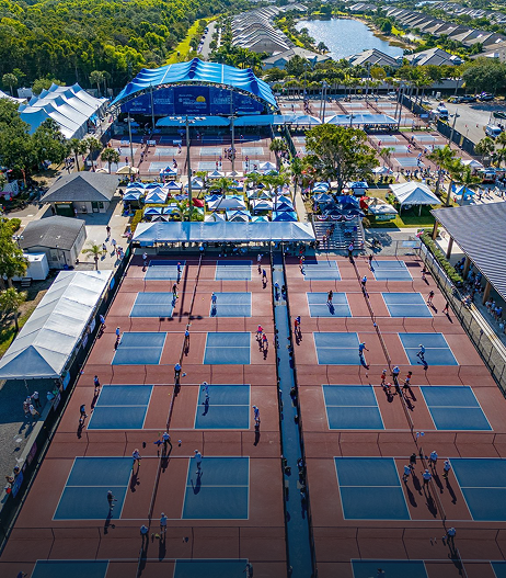 Aerial view of an outdoor pickleball complex with multiple blue courts and a central pool area.