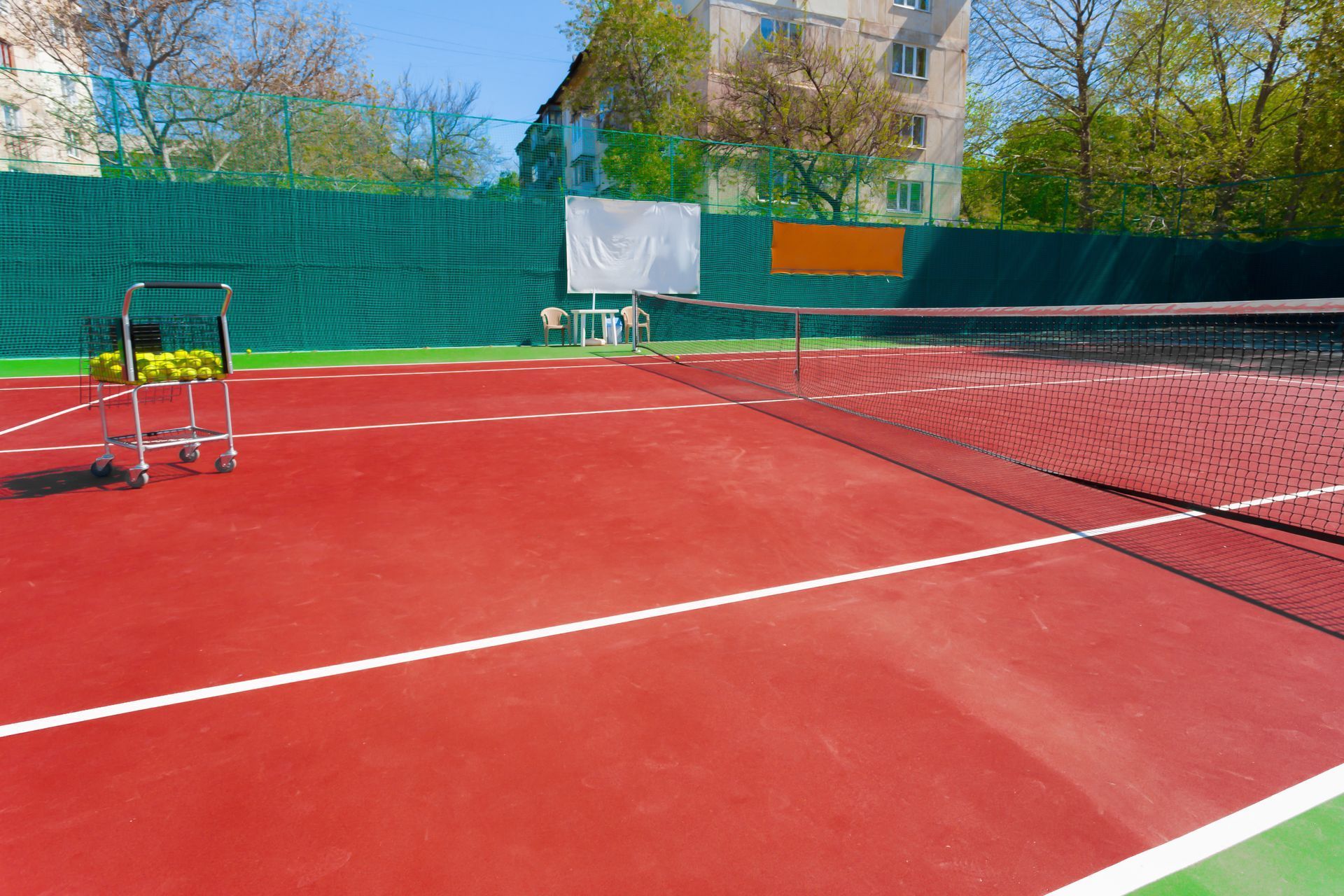 Empty red tennis court with net, ball cart, and green fence under a clear blue sky