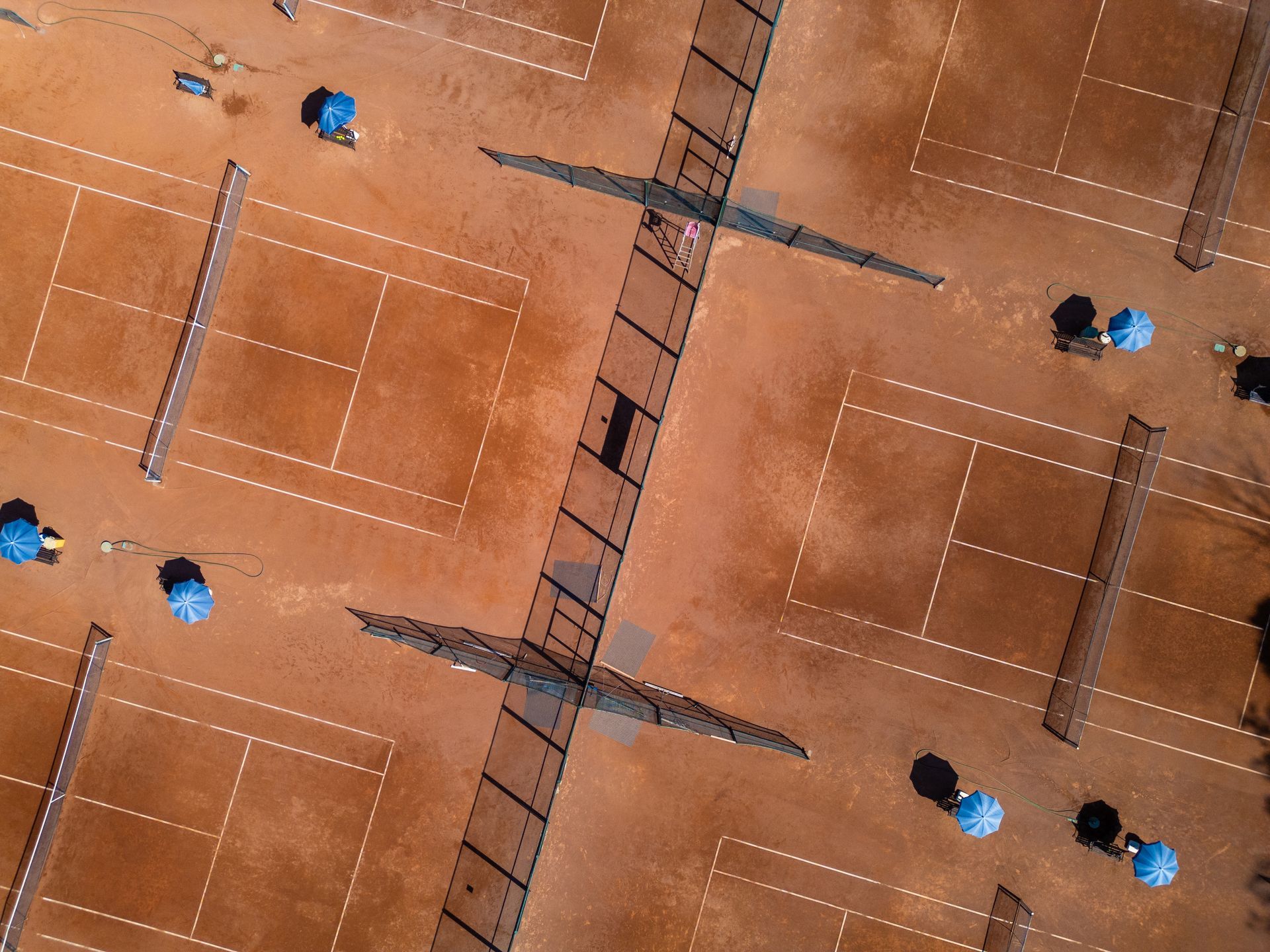 Aerial view of multiple red clay tennis courts lined up outdoors under bright sunlight.