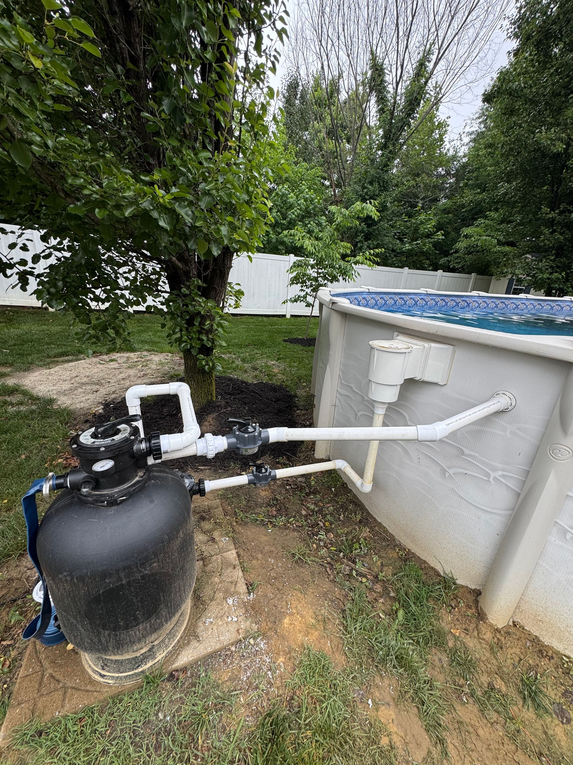 Above-ground pool with black filter pump and white pipes beside a tree in a backyard