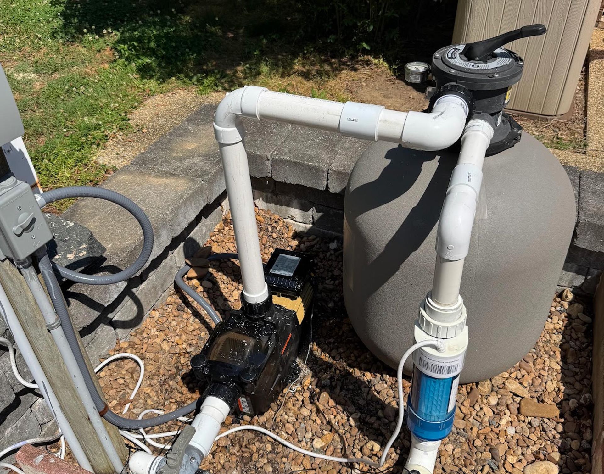 Pool pump and filter setup with white PVC pipes, black pump, and gray tank outdoors on gravel.