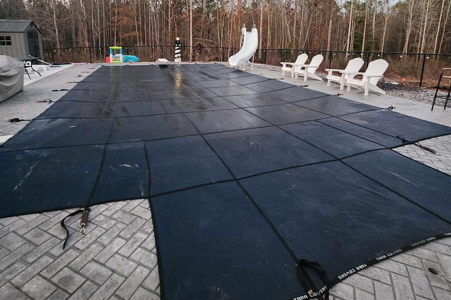Covered backyard pool with black safety cover, white lounge chairs, and trees in the background