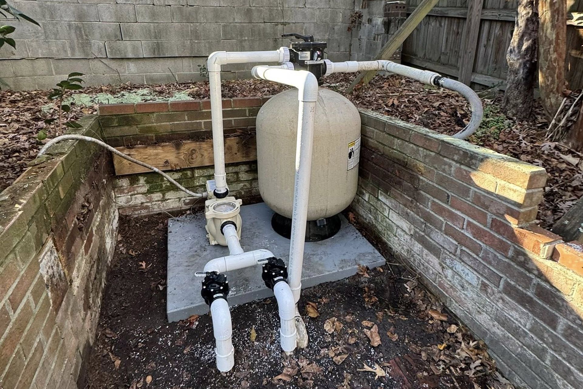 Outdoor pool pump and filter setup with white pipes beside a beige tank in a brick enclosure