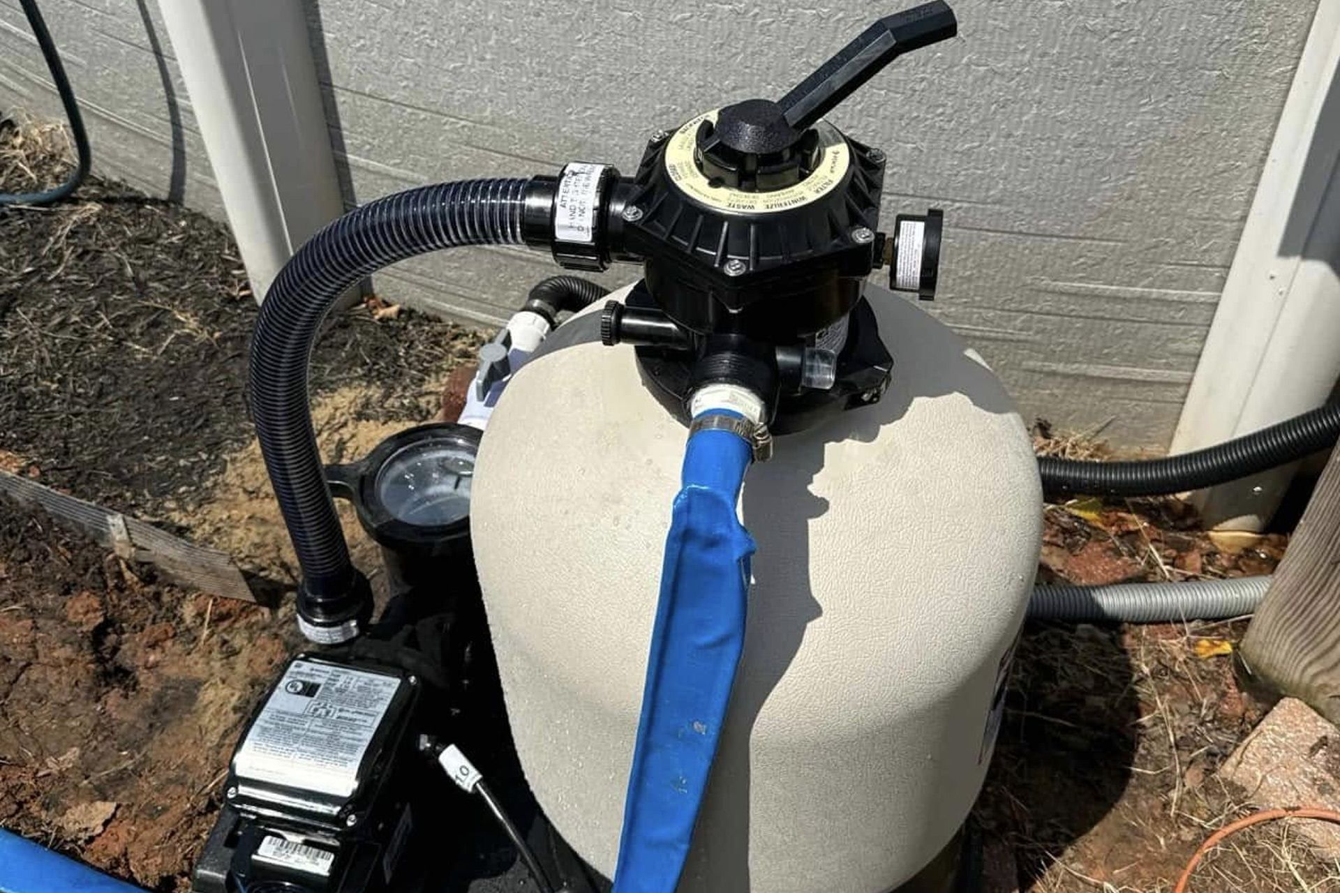 Pool filter tank with a blue hose attached beside outdoor equipment.