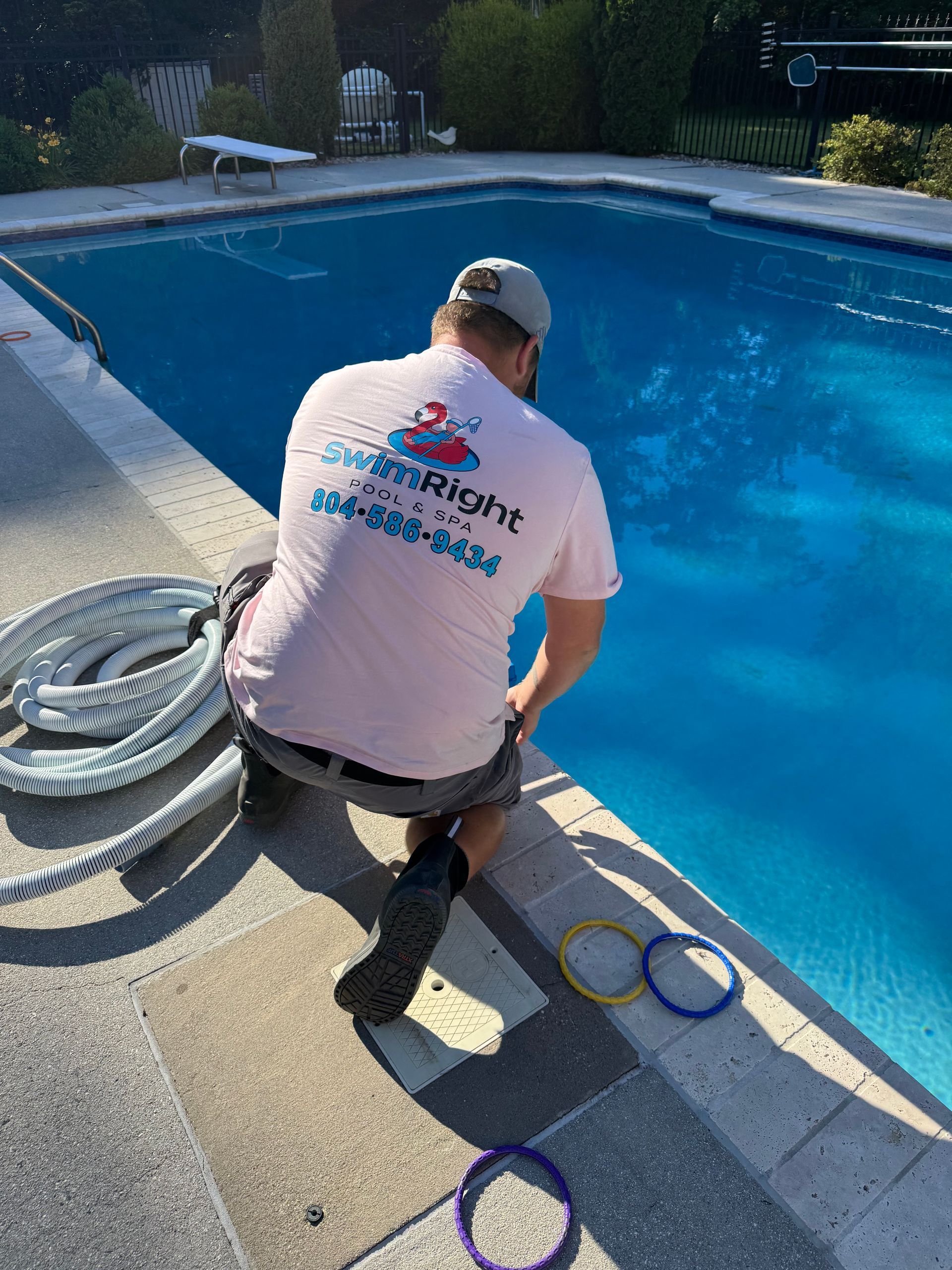 Worker kneeling by a blue swimming pool, handling hoses and cleaning equipment beside the deck.