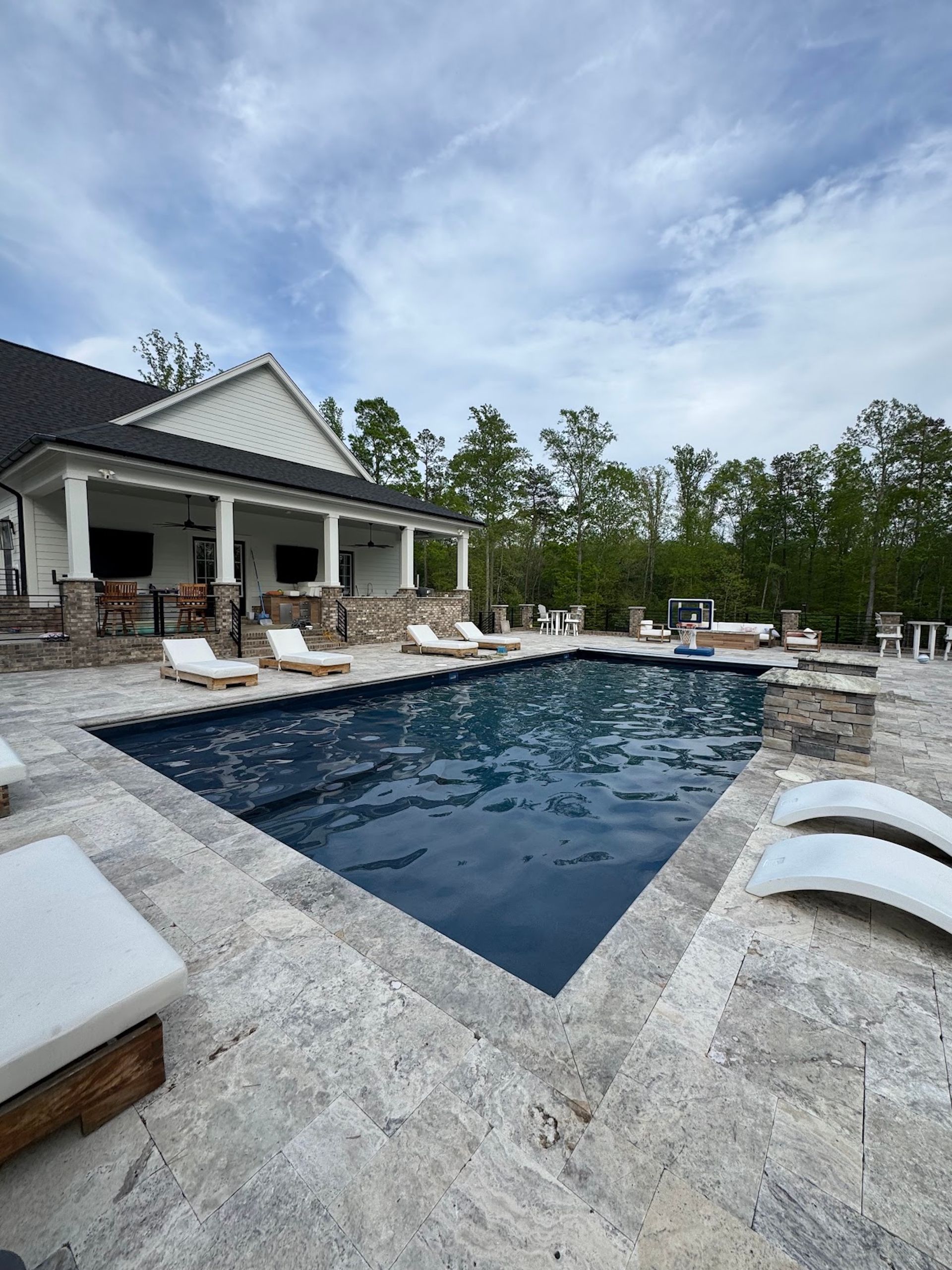 Luxury backyard pool with white loungers, stone patio, and a covered patio under a cloudy sky