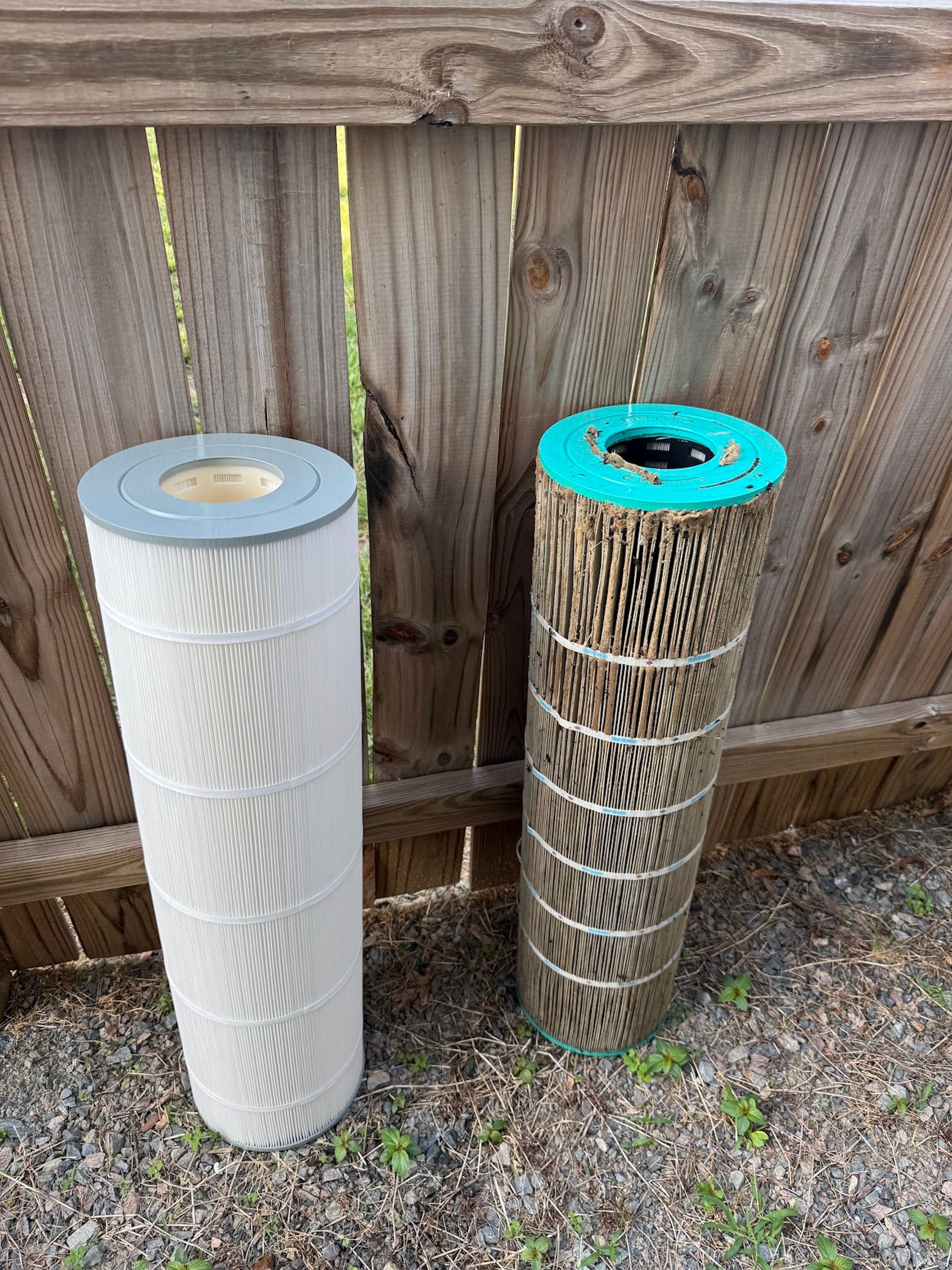 Two cylindrical outdoor air filter cartridges leaning against a wooden fence on gravel.