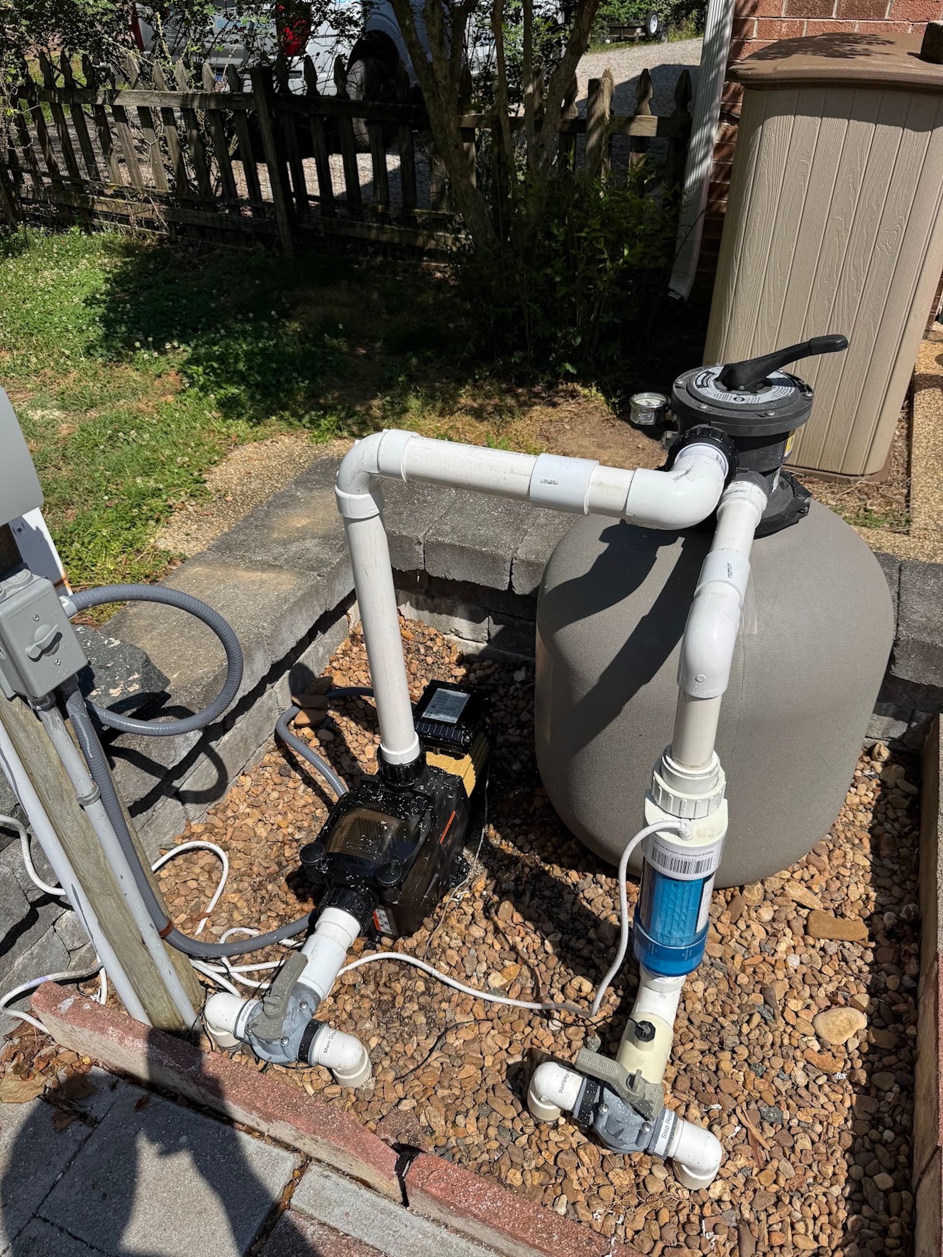 Outdoor pool pump and filter system with white PVC pipes and black motor on gravel beside a fence