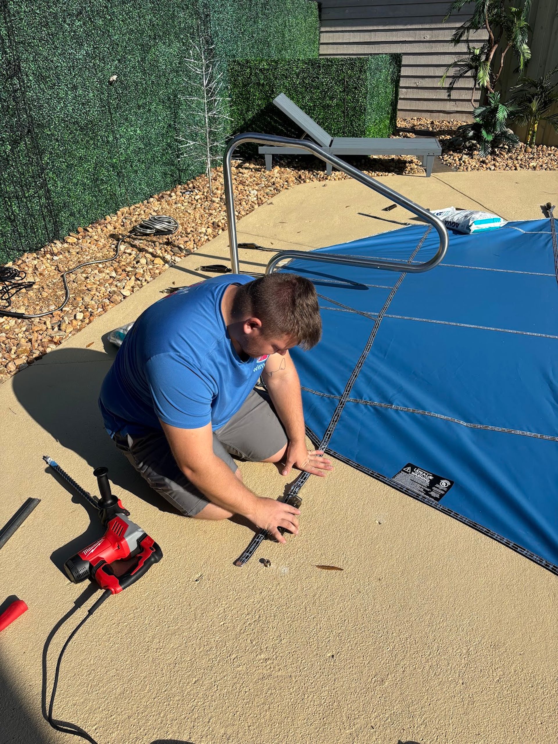 Person securing a blue canopy cover with tools on a gravel patio in sunlight