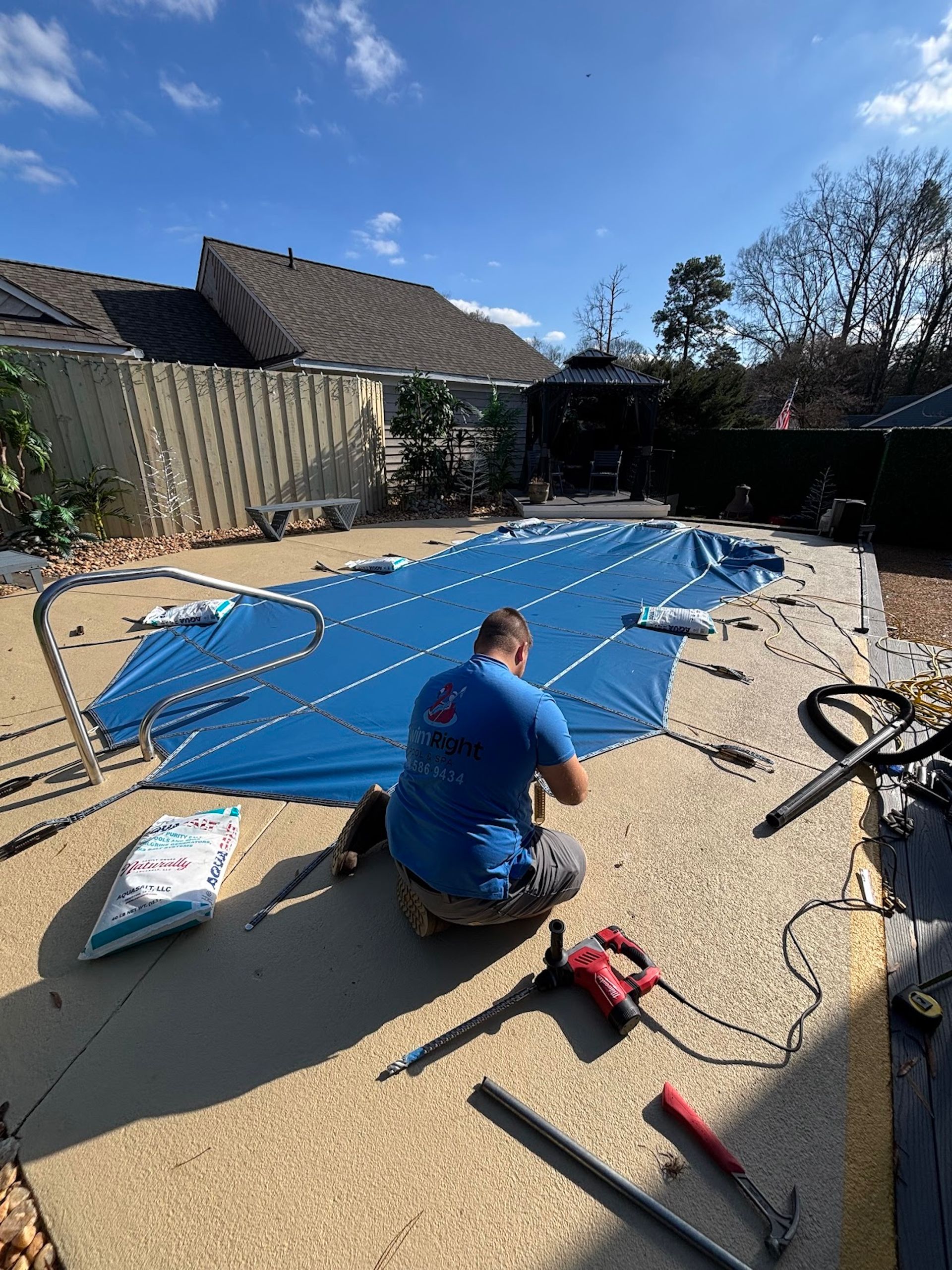 Person securing a blue tarp over a backyard pool with tools on a sunny day