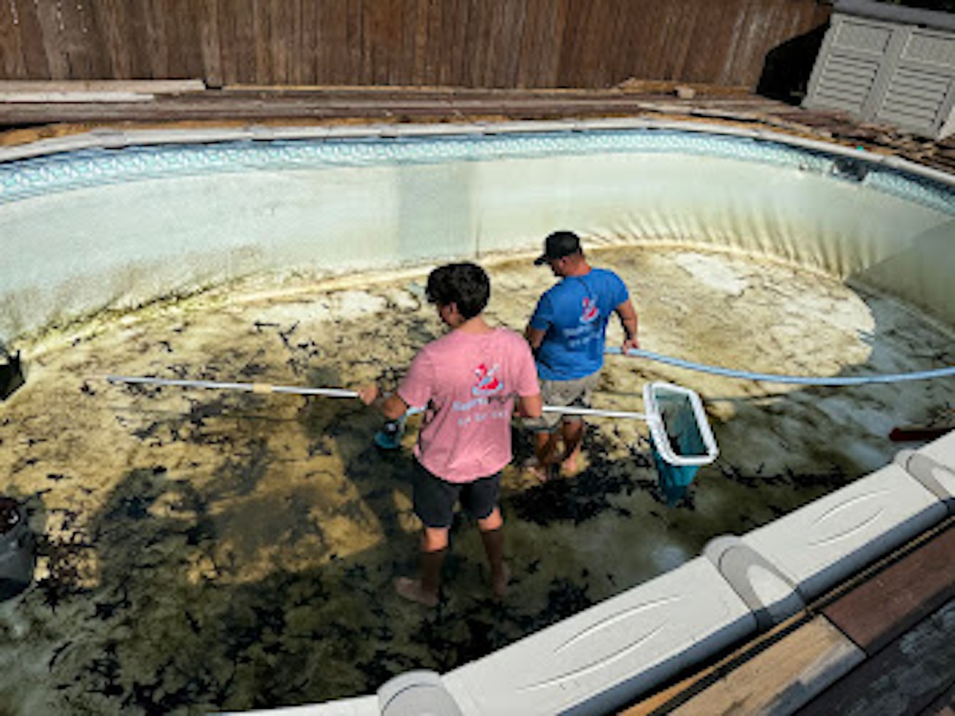 Two workers cleaning a dirty empty swimming pool with a net and hose