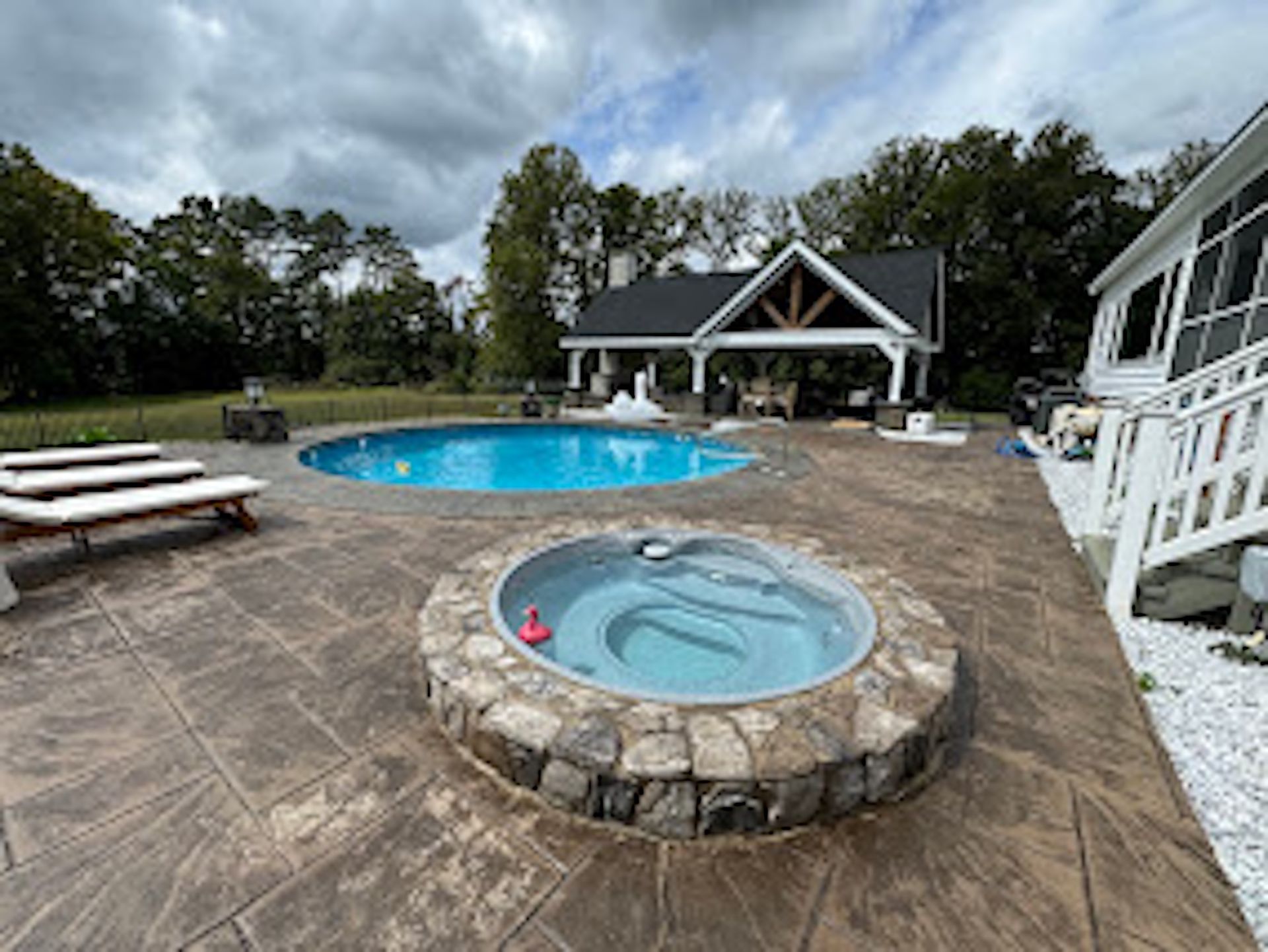 Backyard patio with pool, stone hot tub, lounge chairs, and a covered gazebo under cloudy skies