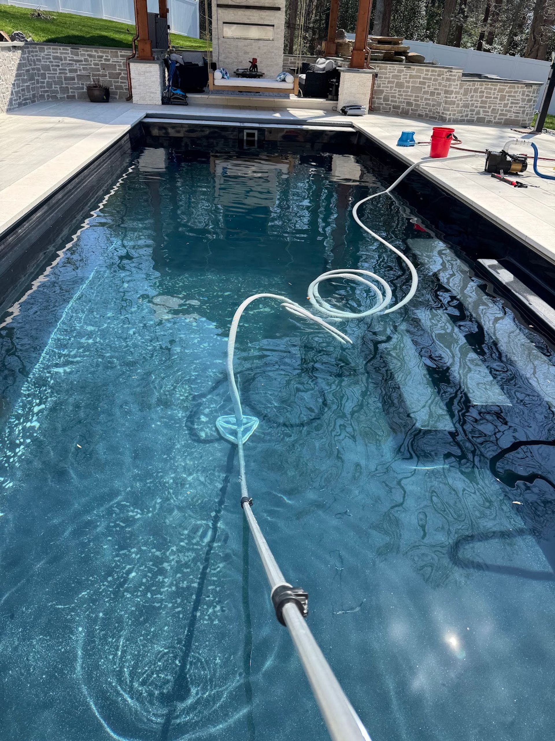 Swimming pool being cleaned with a vacuum hose, water ripples visible and poolside tools nearby