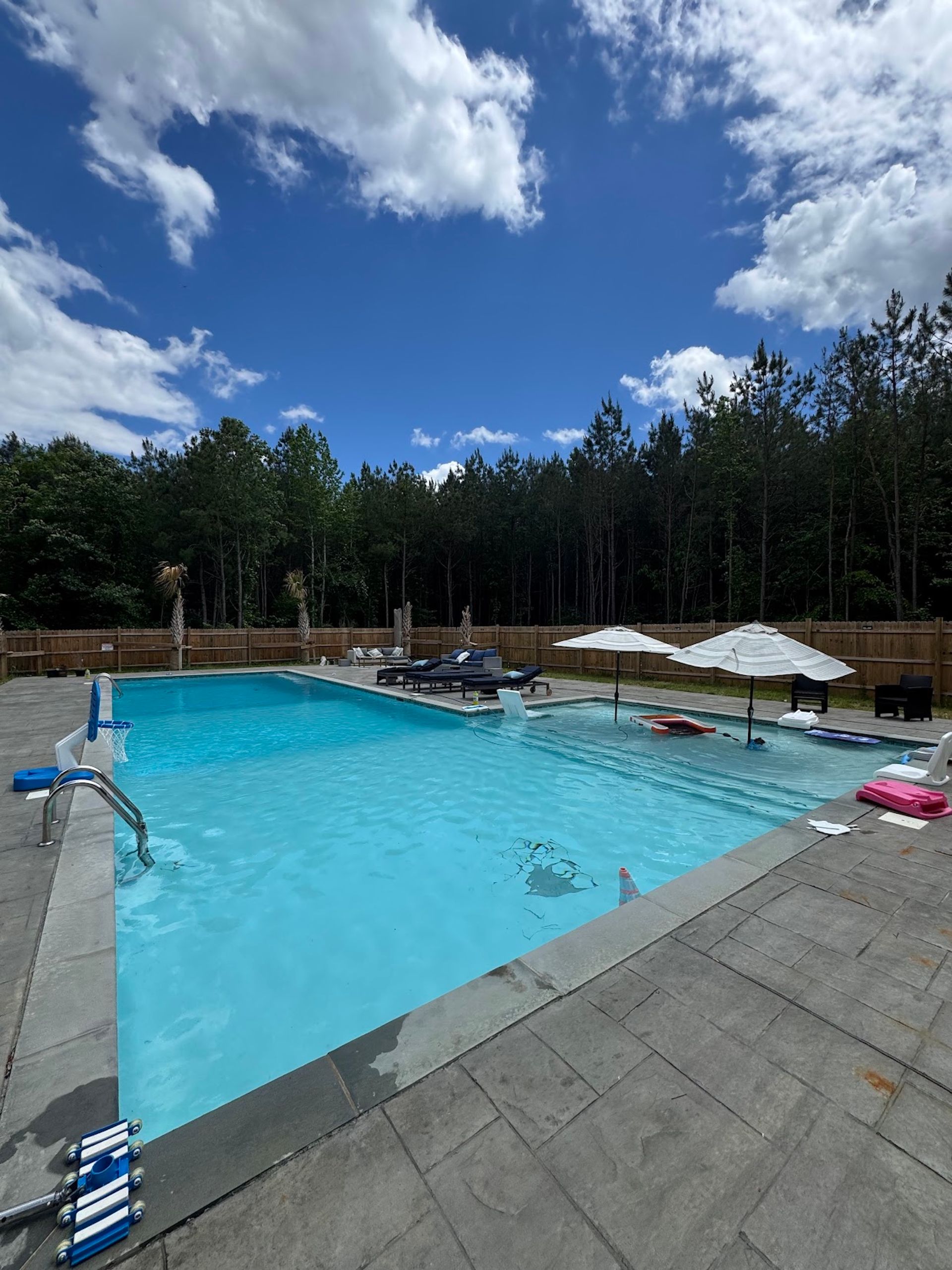 Outdoor swimming pool with clear blue water, lounge chairs, umbrellas, and forested surroundings under a partly cloudy sky