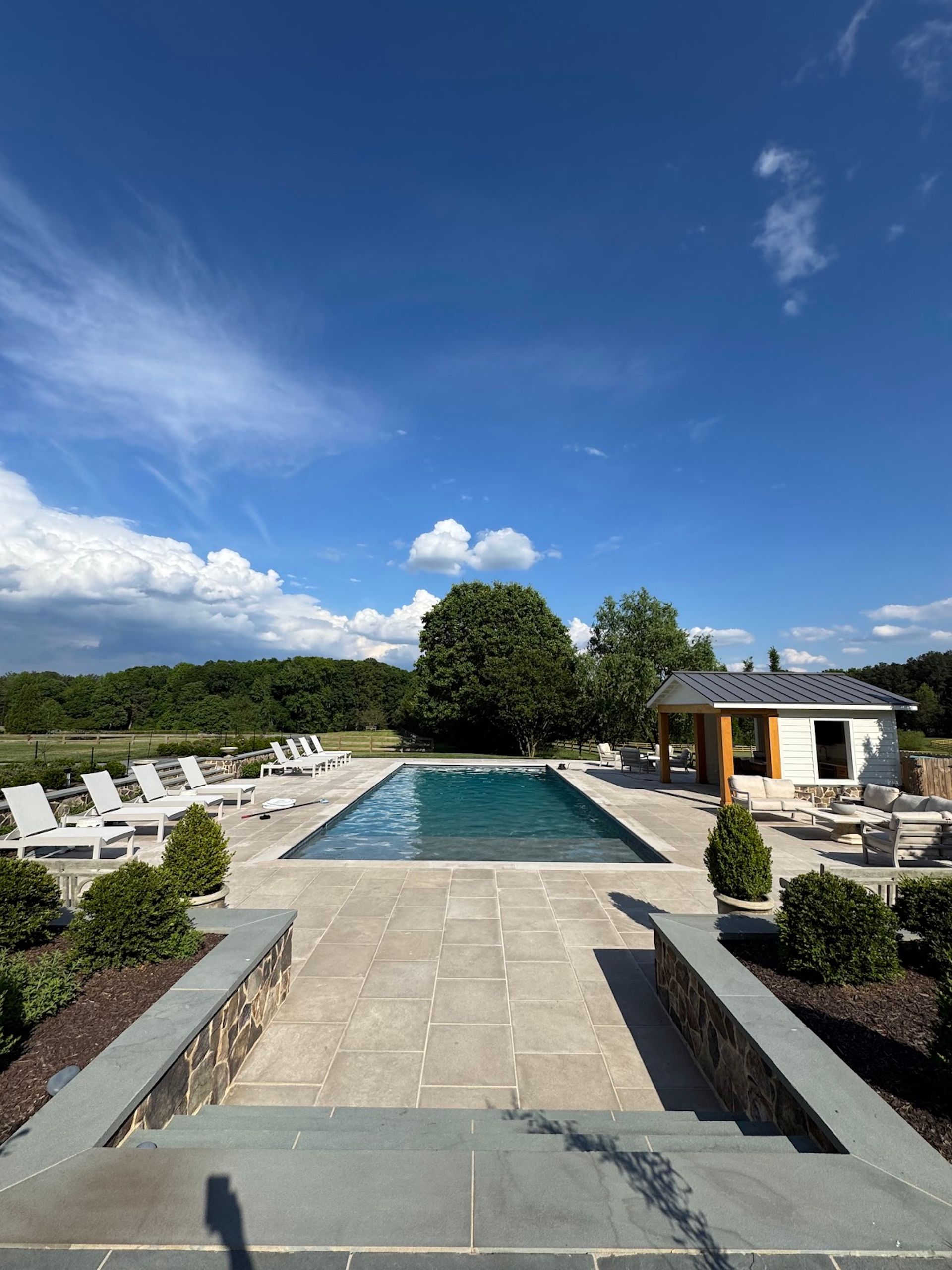 Stone walkway leading to a rectangular outdoor pool under a bright blue sky, with lounge chairs and a pavilion.