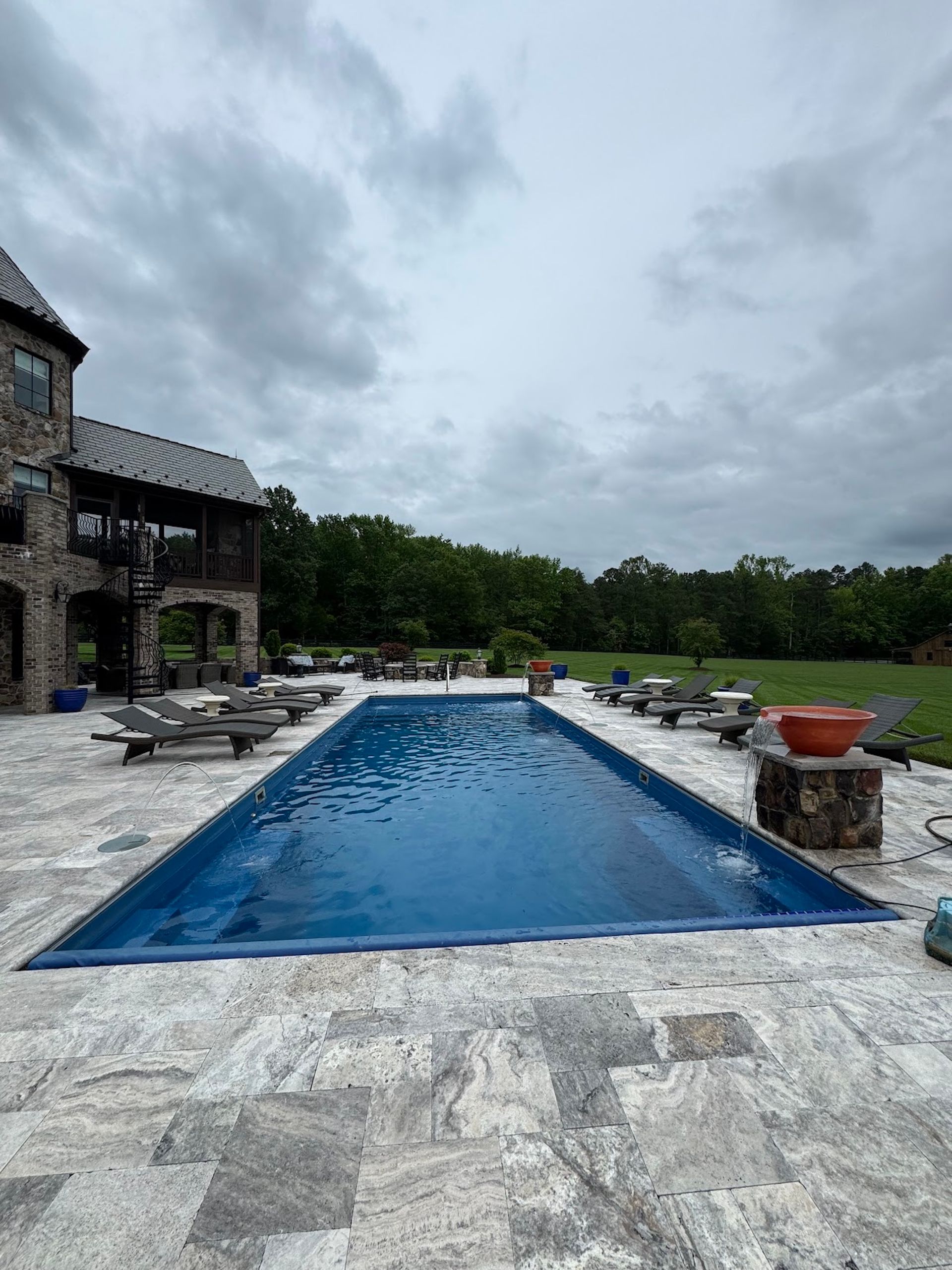 Stone patio with a long blue swimming pool, lounge chairs, and a cloudy sky beside a house