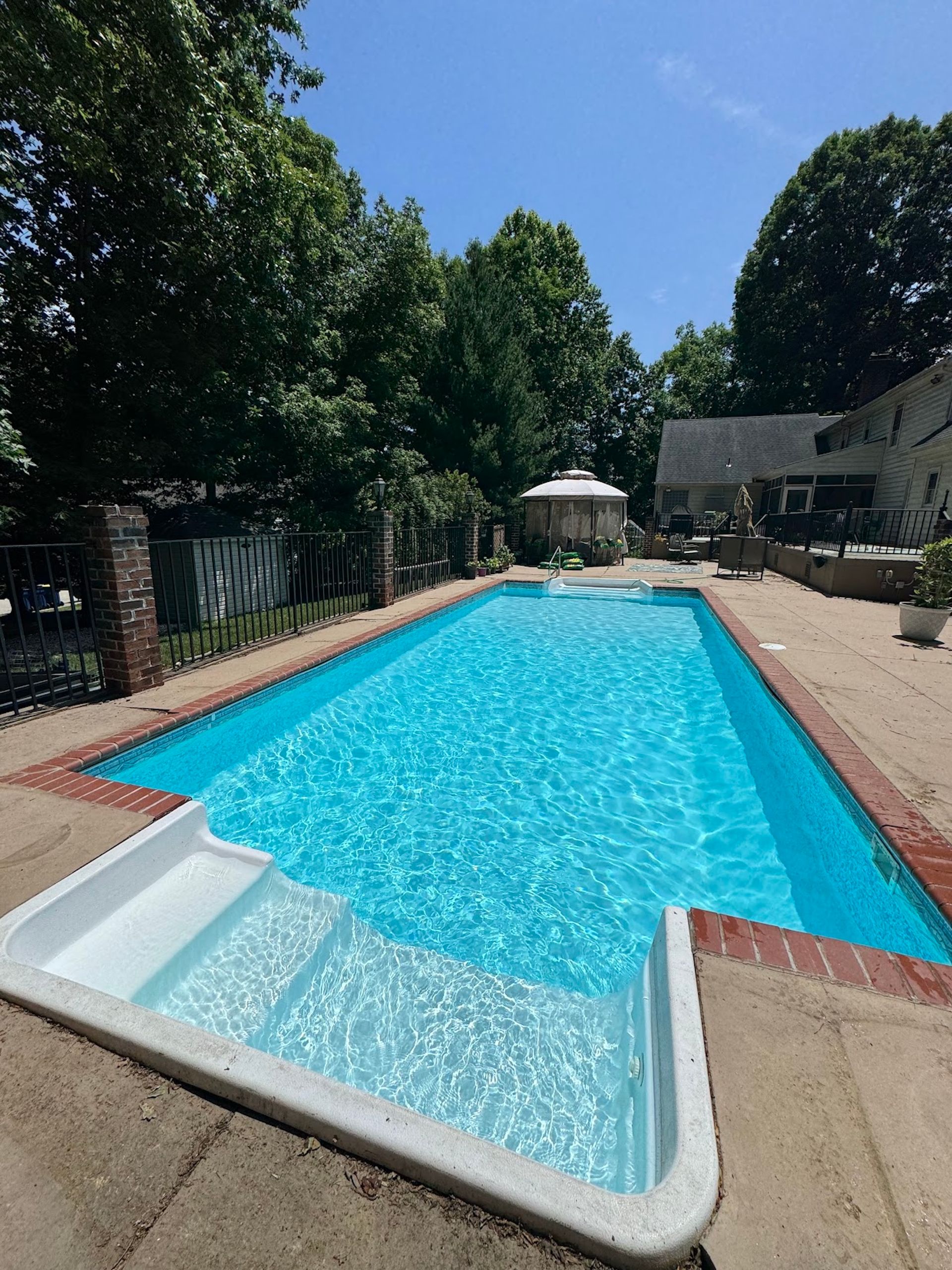 Backyard swimming pool with bright blue water, trees, and patio seating on a sunny day