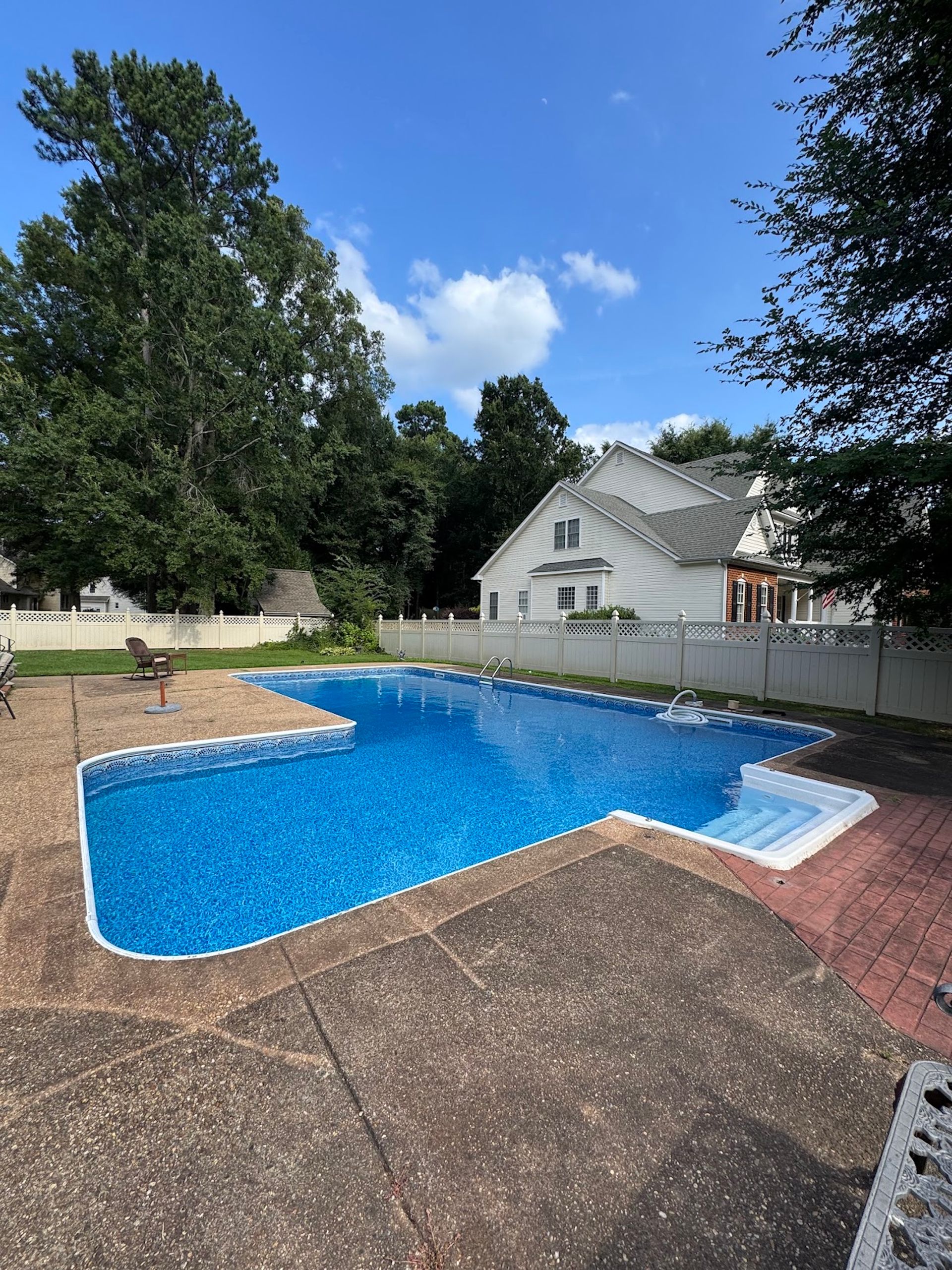 Backyard swimming pool with blue water, white fence, trees, and a house in the background