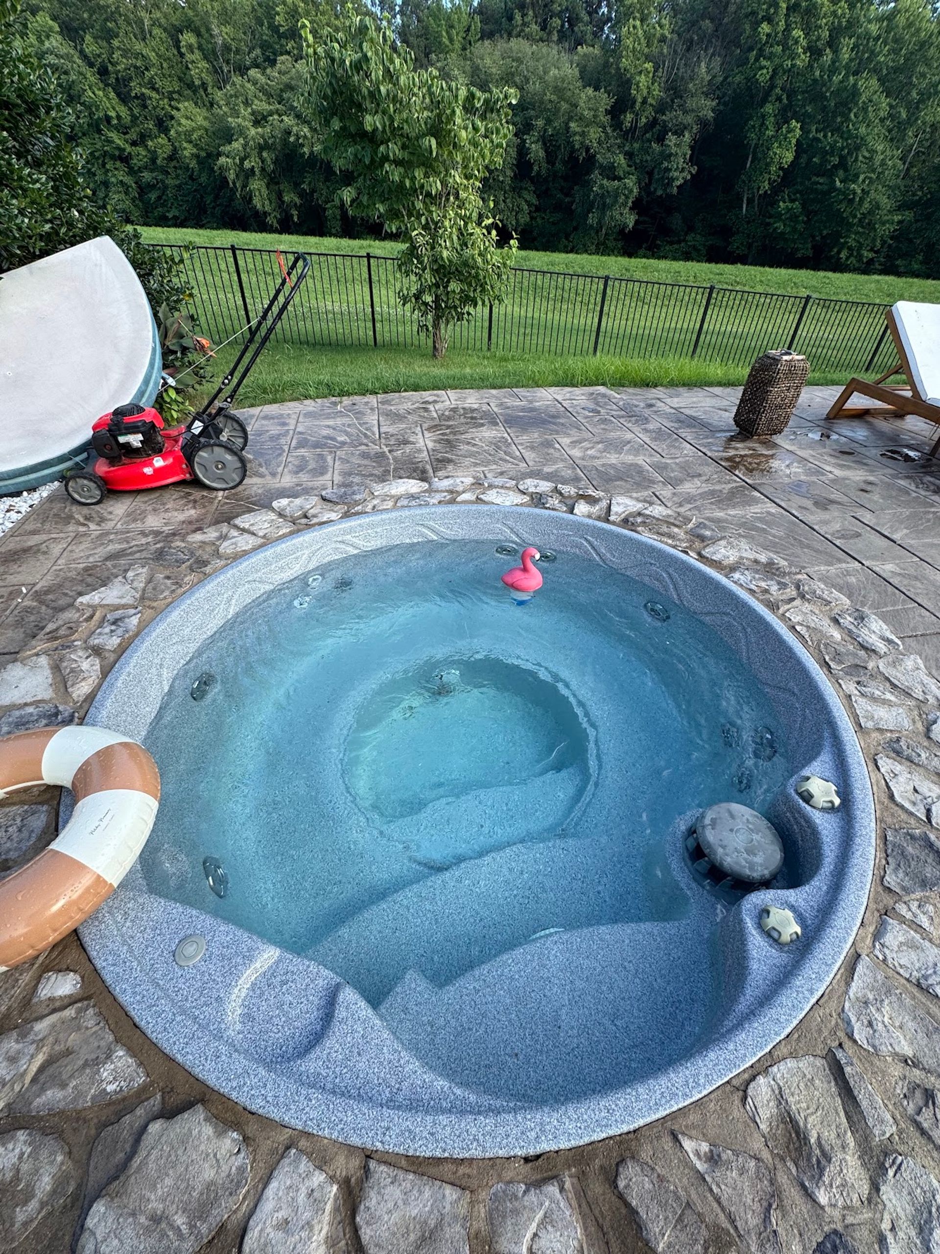 Empty backyard hot tub with blue water beside a stone patio and green fence, surrounded by garden and chairs