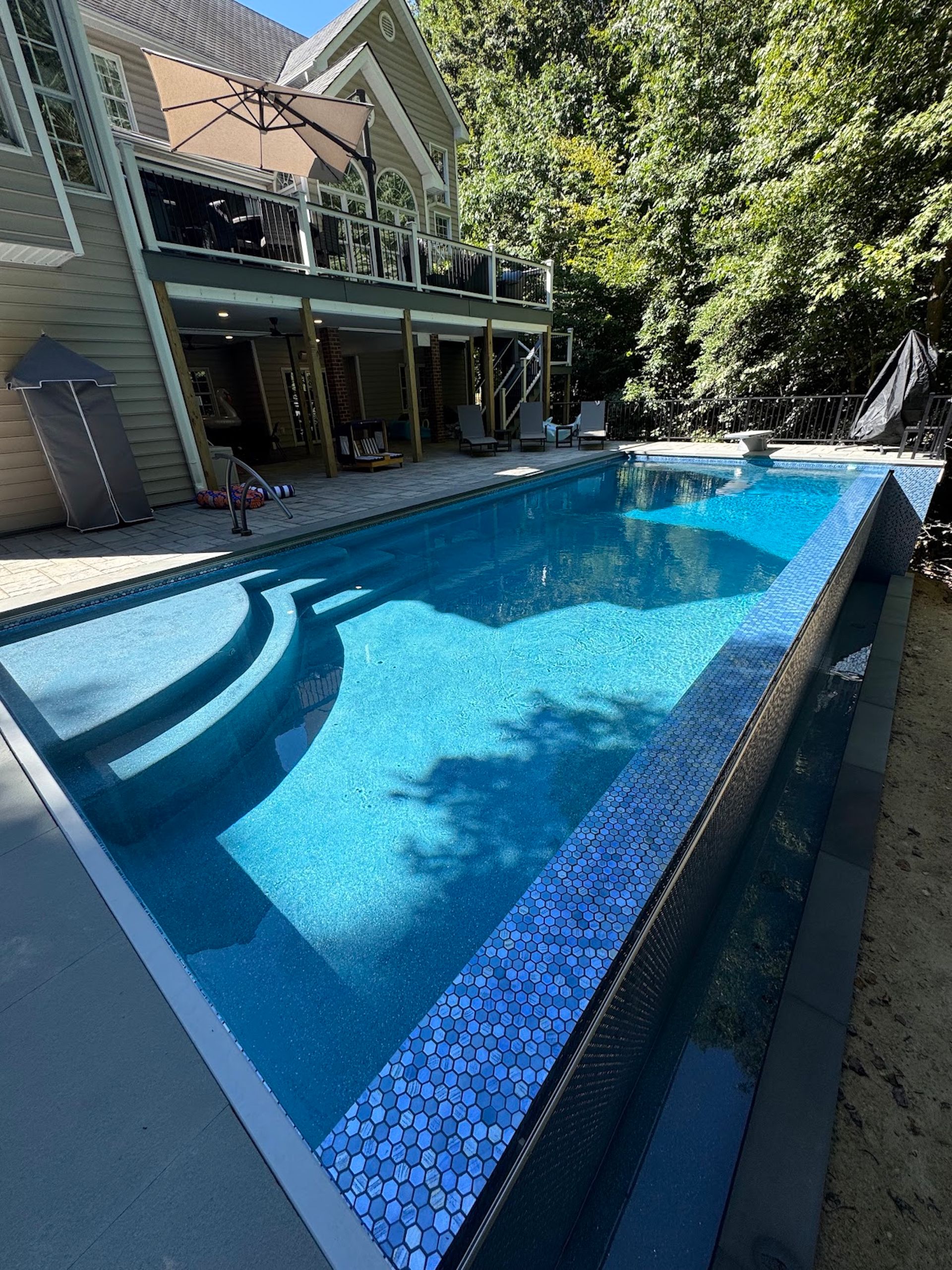 Long blue lap pool beside a modern house, with patio and trees in bright sunlight