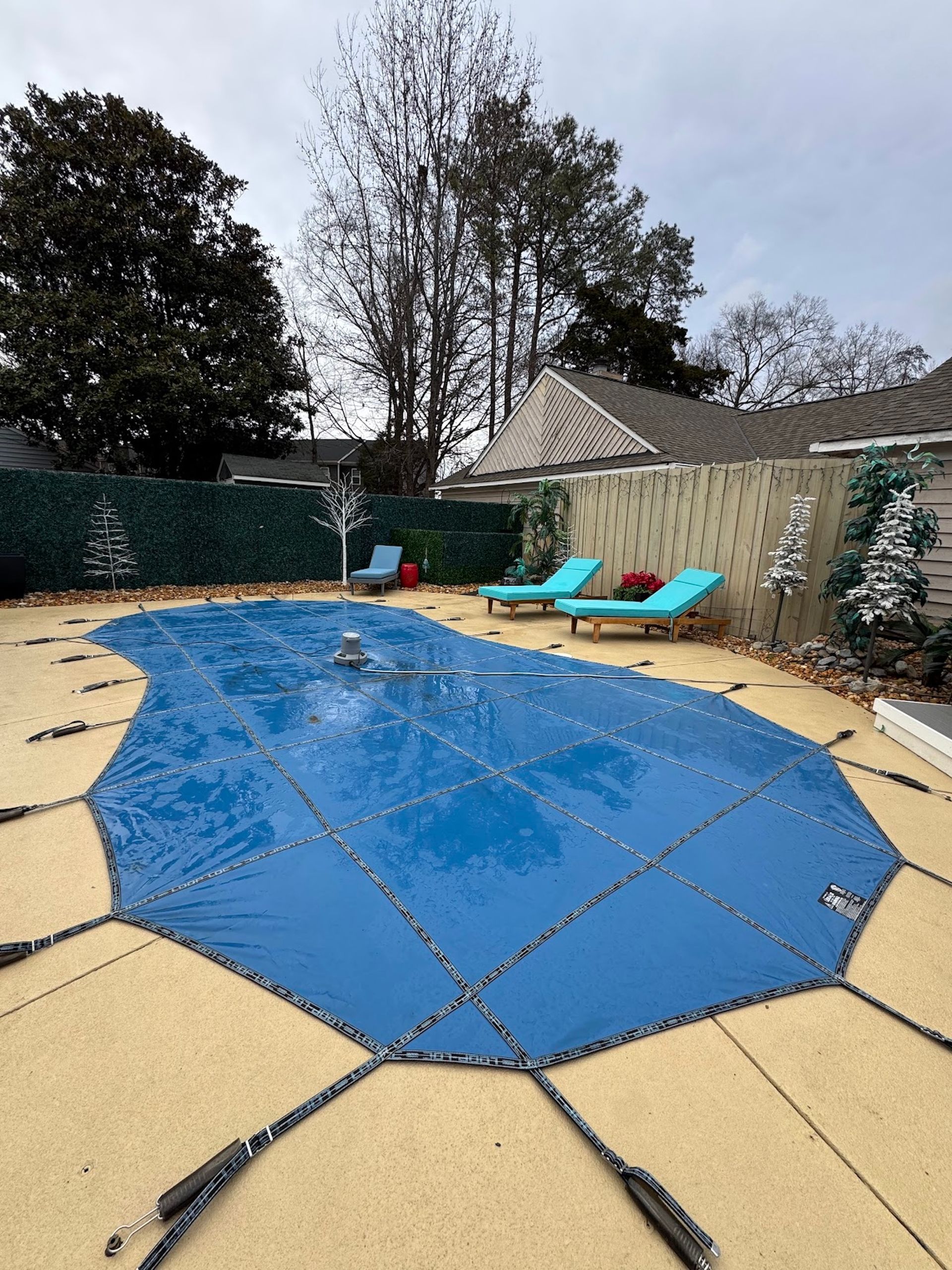 Backyard pool covered with a blue tarp, surrounded by tan pavers, lounge chairs, and trees