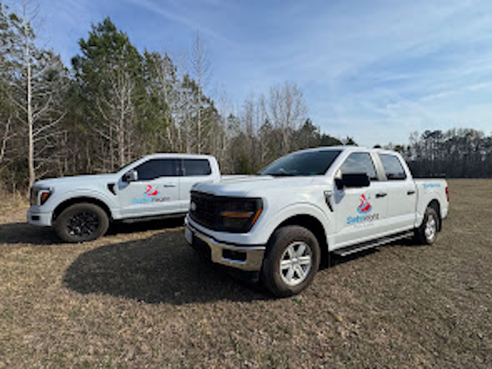 Two white pickup trucks parked on grass in a wooded area under a clear sky