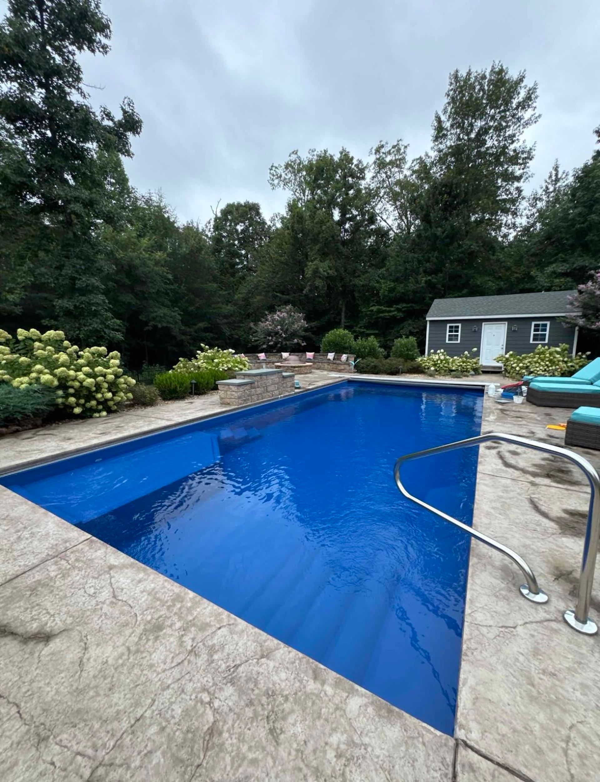 Backyard swimming pool with blue water, stone deck, and patio chairs beside a small shed.