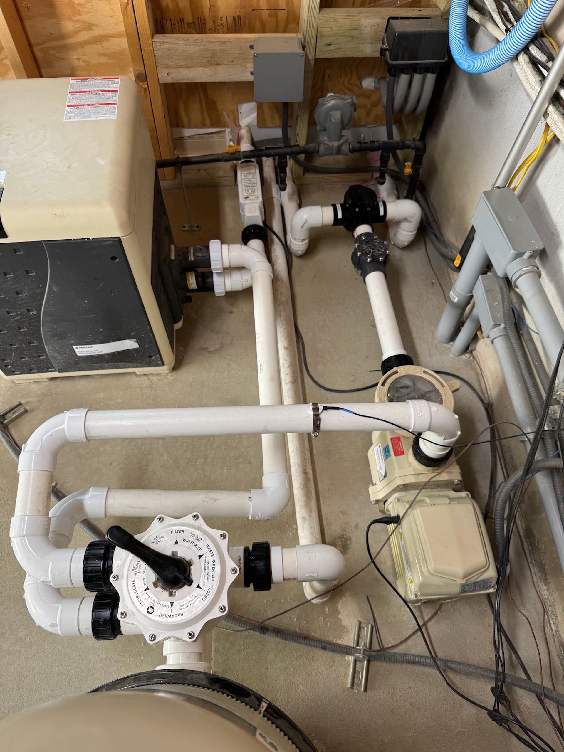 Basement pool plumbing with white PVC pipes, pump, heater, and electrical wiring on concrete floor