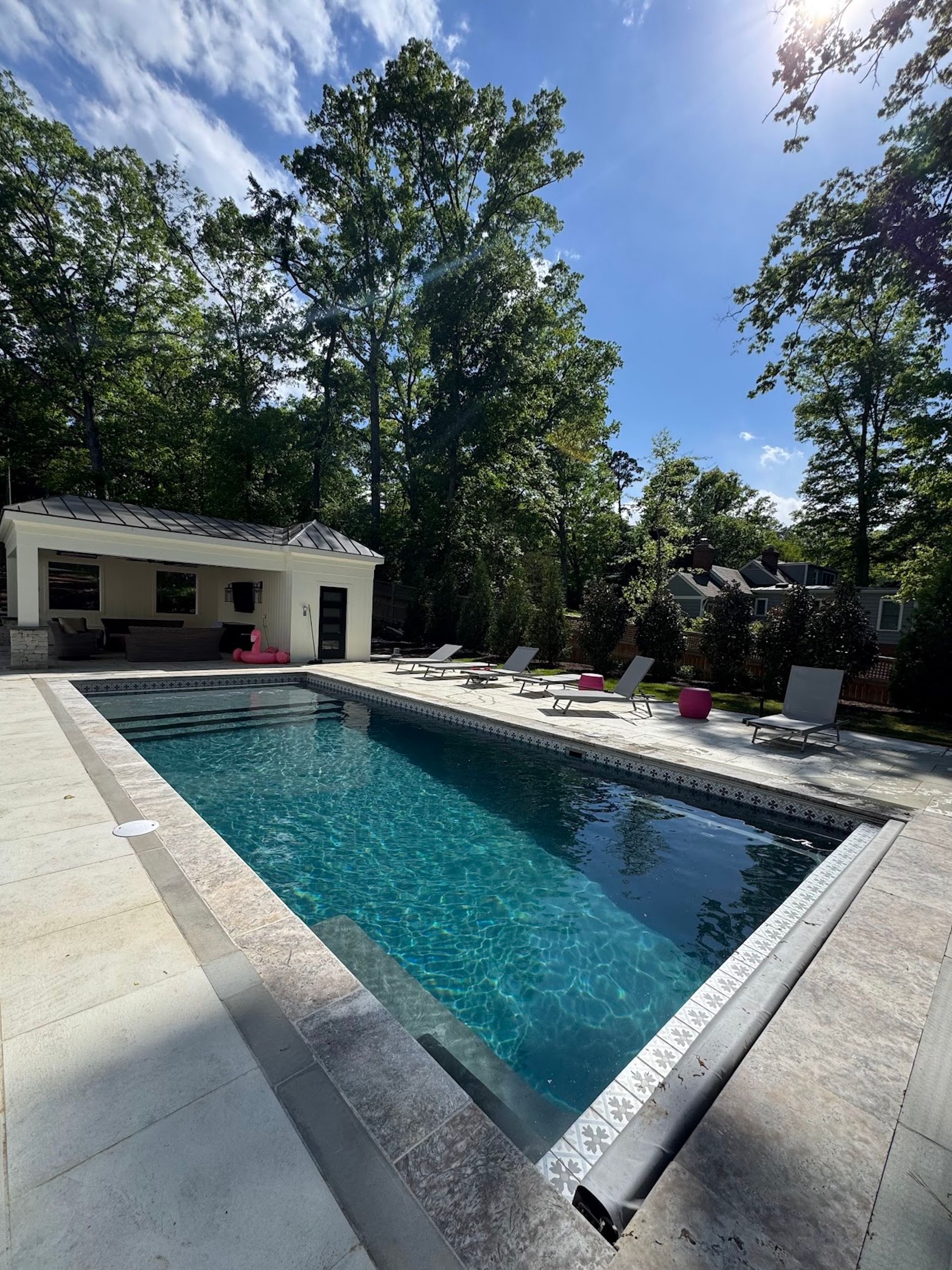 Backyard pool with turquoise water, white stone deck, and trees under a bright blue sky