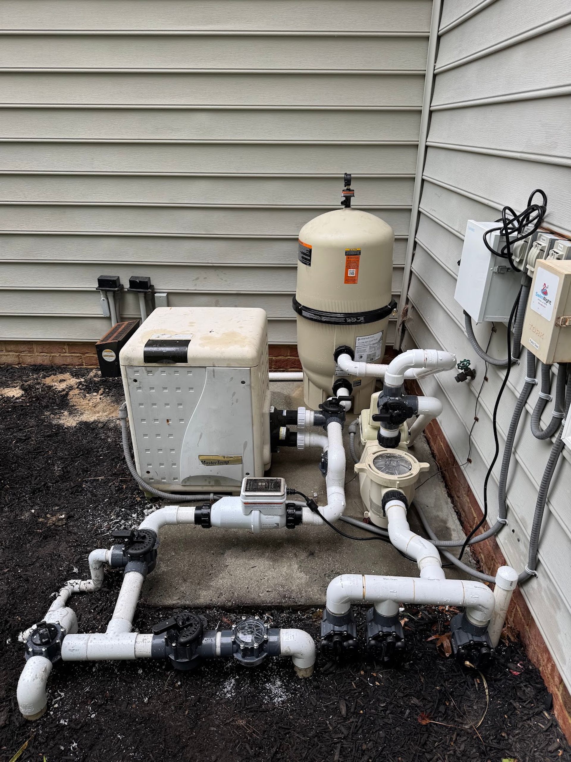 Pool equipment beside house wall with pipes, pump, filter tank, and electrical unit on gravel.