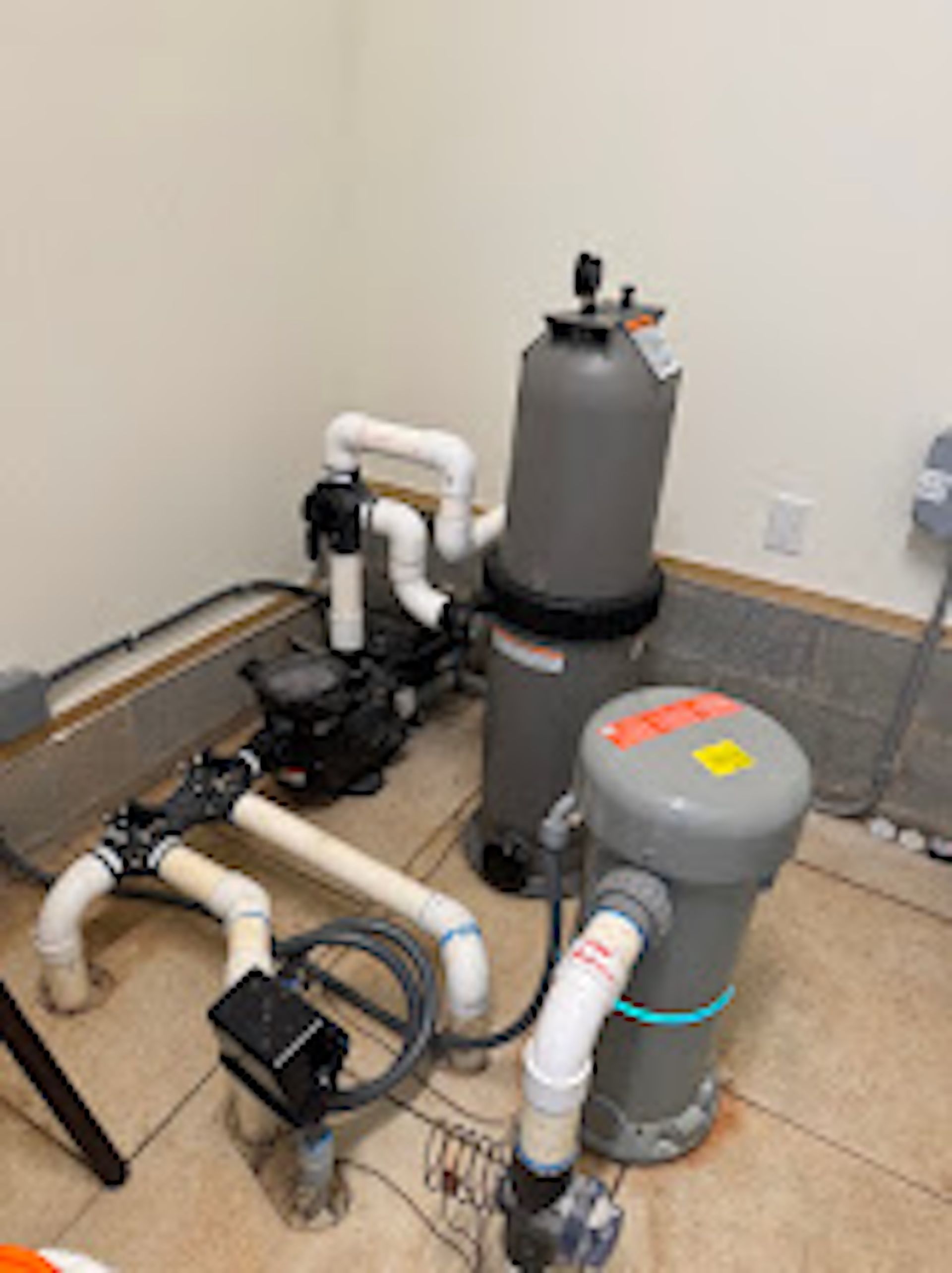 Pool pump and filter equipment with white pipes in a utility corner on a tiled floor.
