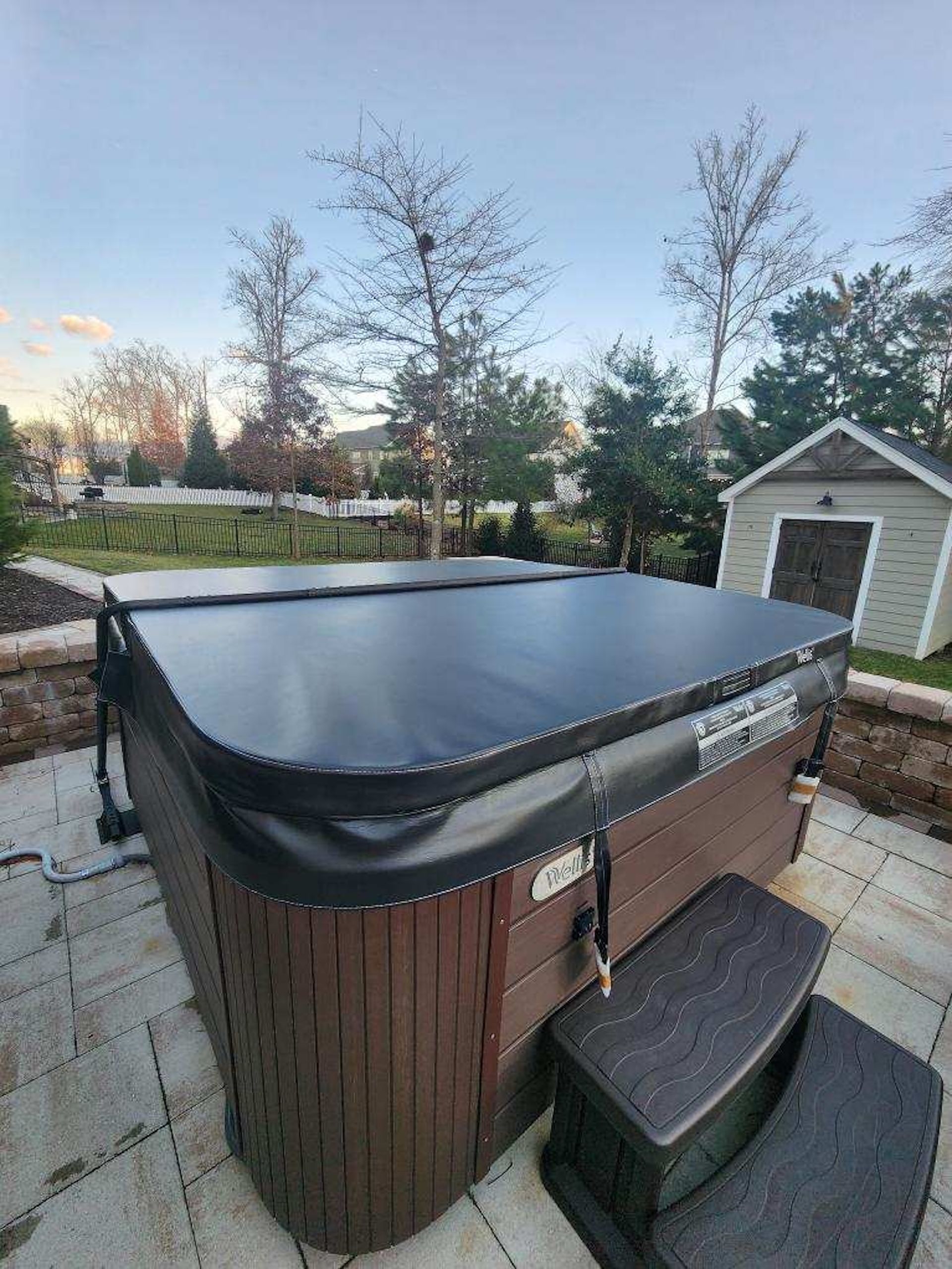 Covered outdoor hot tub on a patio beside a small shed at dusk