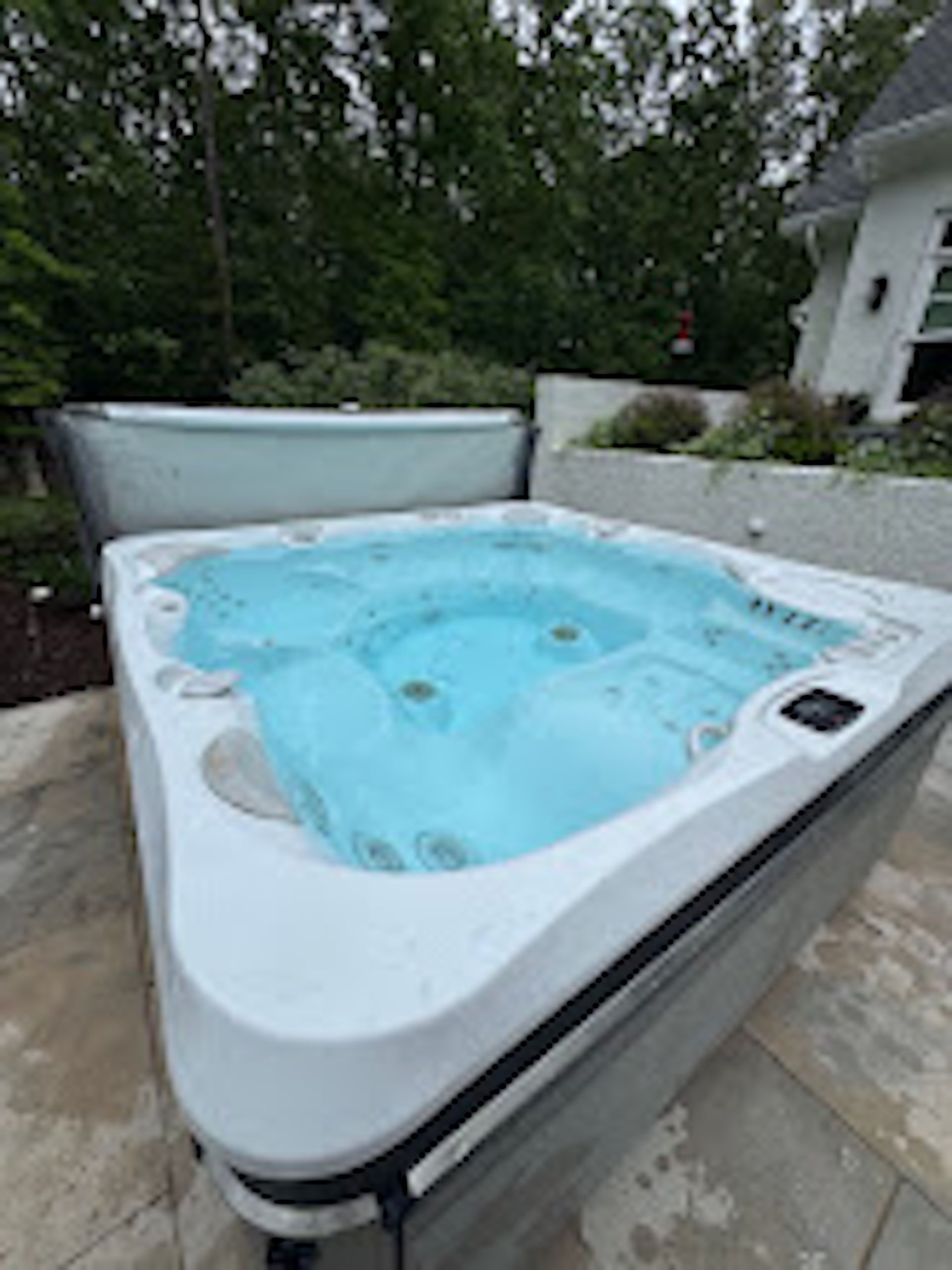 White hot tub with blue water on a stone patio, surrounded by greenery outdoors.