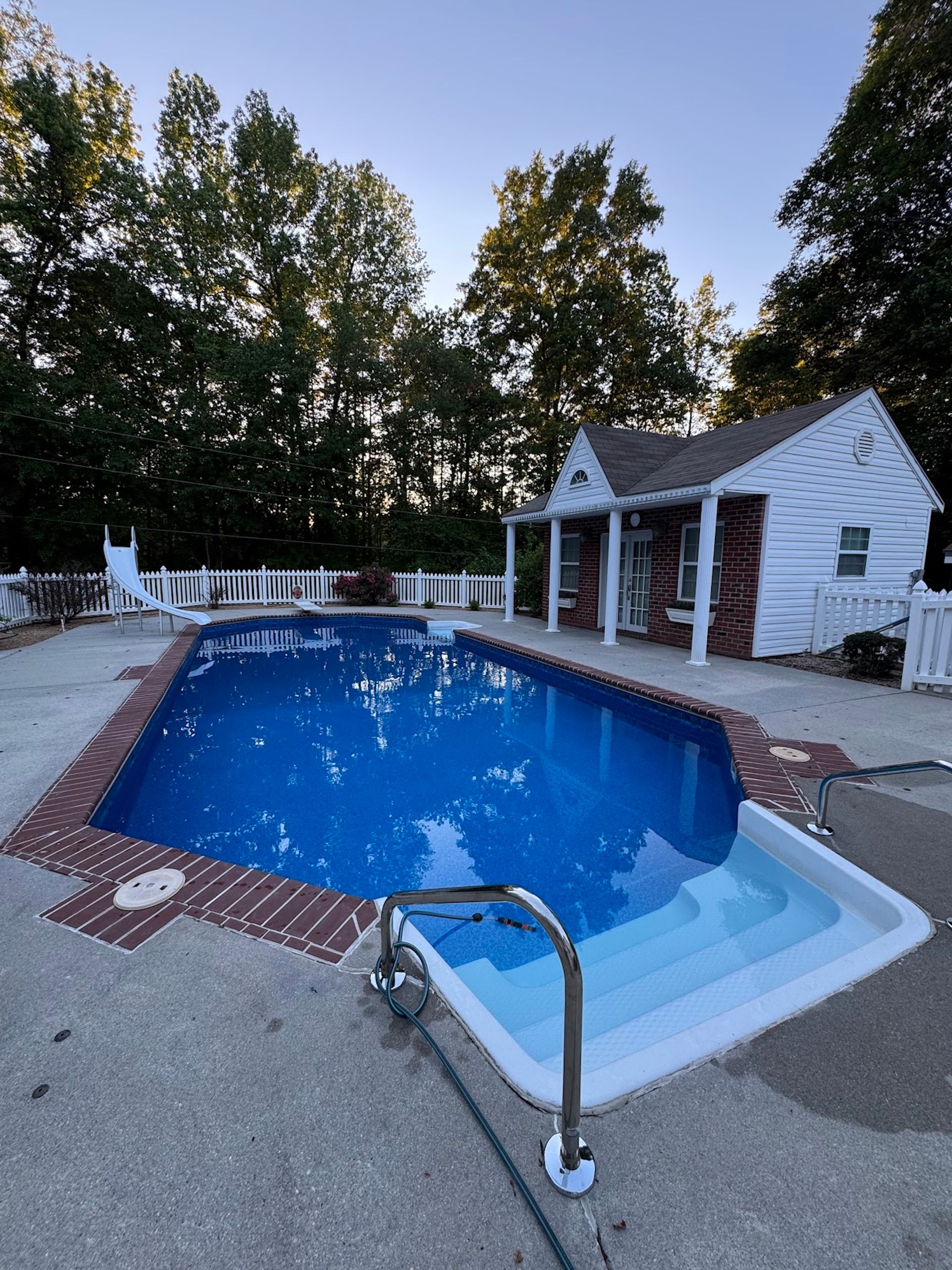 Backyard pool with blue water, brick coping, and a small white pool house at dusk
